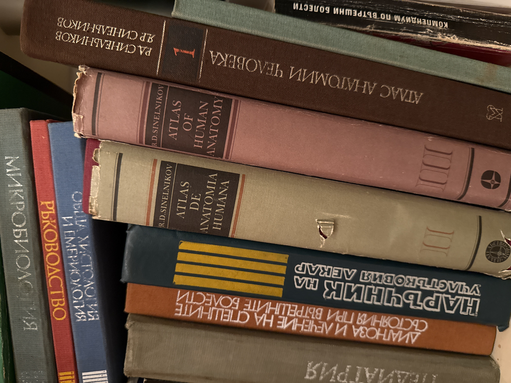 The image is a close-up, overhead view of a shelf of books. The books are mostly hardbound and are arranged vertically, leaning slightly. Several of the books have visible titles in Cyrillic script, indicating they are likely in a Slavic language. Some of the titles appear to be medical or anatomical in nature, such as "ATLAS OF HUMAN ANATOMY" which is visible in English on one of the books. The lighting suggests an indoor setting, possibly a library or personal collection. There are no people or animals present in the image. The environment appears to be a bookshelf or a similar storage area for books. The city of Targovishte, Bulgaria is not visible in the image and there is no information in the image to suggest its location there.