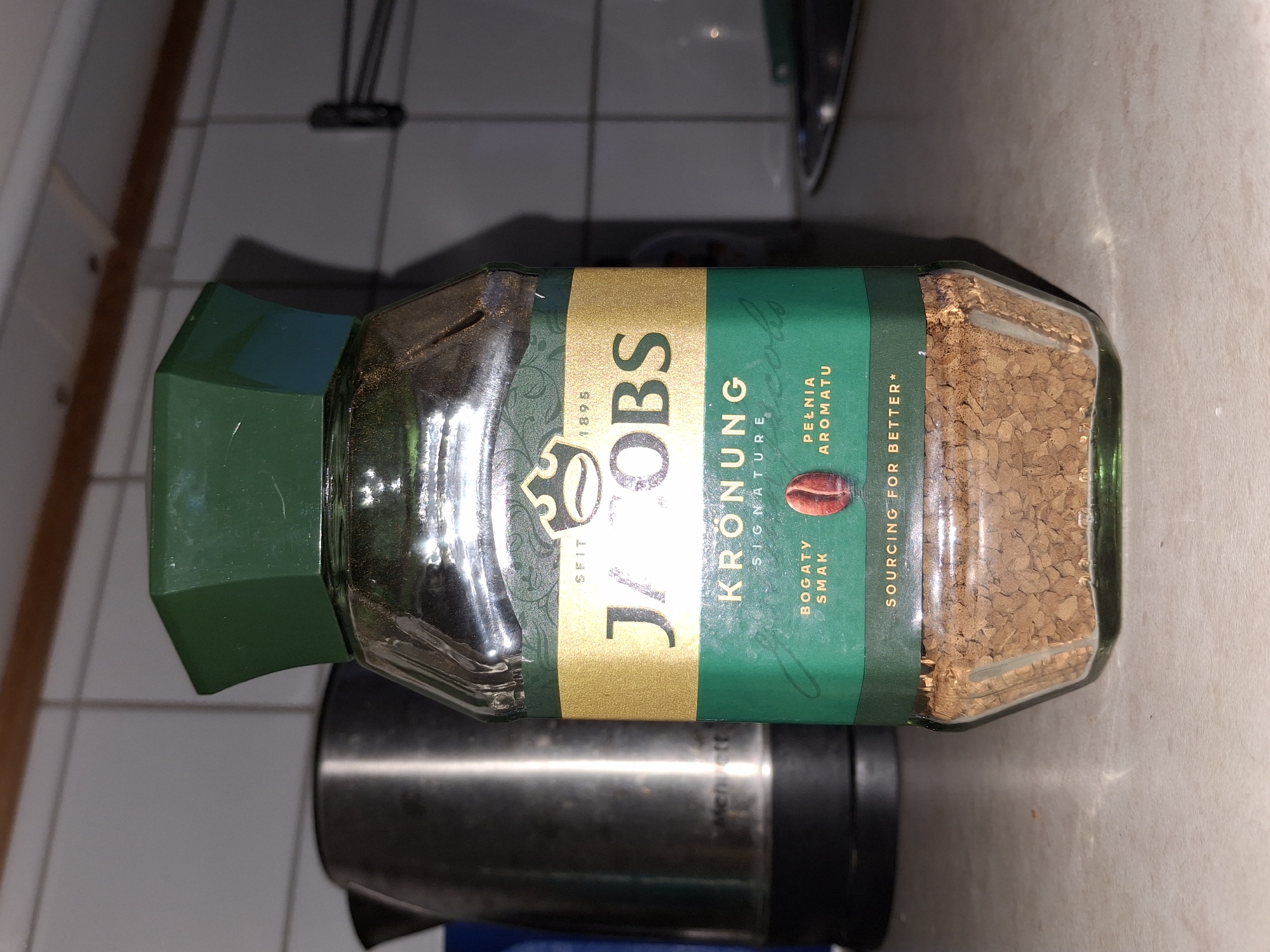 A jar of Jacobs Kronung instant coffee is placed on a countertop, likely in a kitchen setting in Åsgårdstrand, Norway. The jar is predominantly green and gold, with the brand name "JACOBS" prominently displayed in gold lettering. The coffee inside is visible at the bottom of the glass jar, appearing as small brown granules. To the left of the coffee jar, a silver electric kettle is partially visible. The background consists of tiled walls and a wooden surface, suggesting a domestic environment. The lighting in the scene is somewhat dim, with shadows cast on the countertop.