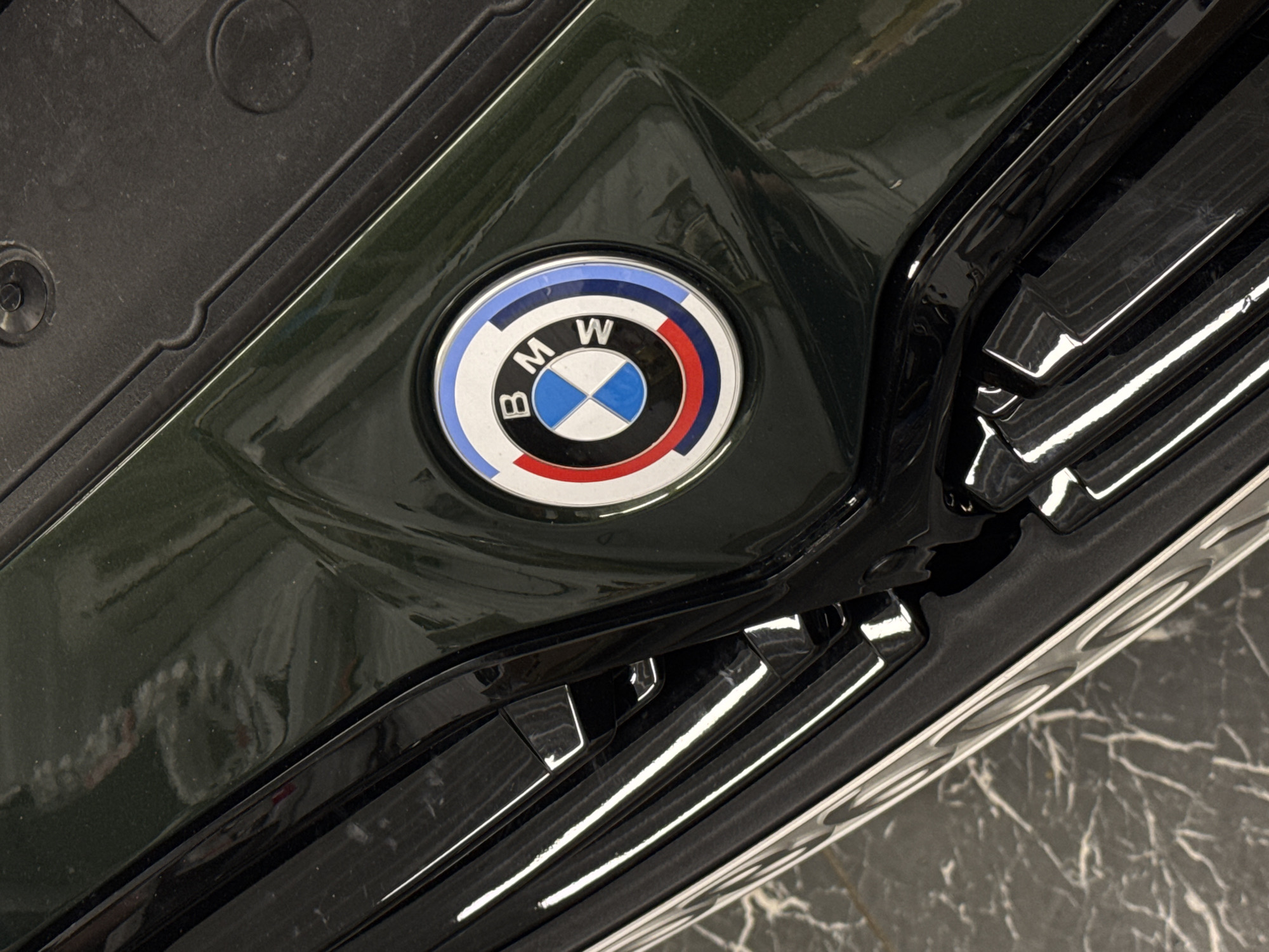 The image displays a close-up, downward-angled view of the front section of a dark green (possibly British Racing Green) vehicle. Prominently featured in the upper-center is a circular BMW emblem, specifically the "50 Years of BMW M" version, which includes the standard blue and white quartered inner circle surrounded by an outer ring segmented into blue, dark blue, and red. To the left and below the emblem, a portion of the vehicle's black grille with vertical slats is visible, reflecting white light sources. Above the emblem, a textured black plastic panel, likely part of the engine bay cover or cowl, extends to the upper right. The paintwork exhibits a high gloss finish with visible reflections. The immediate environment beneath the vehicle's front is a dark, reflective floor surface with light, irregular veining patterns, consistent with marble or polished stone tiles. No individuals or specific actions are depicted. The exact location within Chaikovsky, Russia, cannot be determined from the visual information provided.