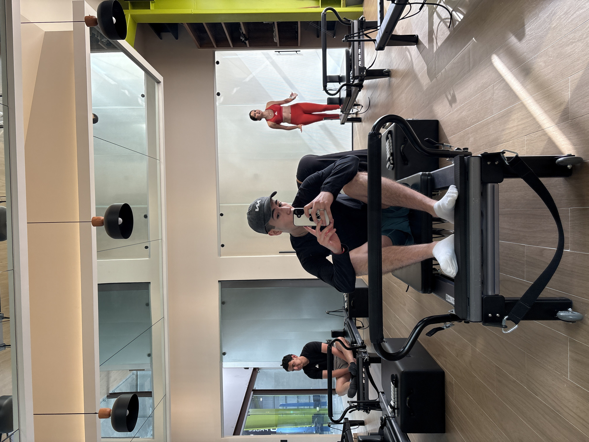 A young man is positioned on a black Pilates reformer, taking a mirror selfie in a modern fitness studio located in San Bernardino Tlaxcalancingo, Mexico. He is wearing a black long-sleeved shirt, dark shorts, white socks, and a black baseball cap with a white "W" logo. He holds a white iPhone with a distinctive graphic, possibly a panda face, on its case.
The mirror reflects not only the man from a different angle but also other occupants of the studio. In the background reflection, a woman in a bright red athletic two-piece set, consisting of a sports bra and leggings, is visible, with a faint "adidas" logo on her top. Further back and to the left in the reflection, another young man is seen sitting on a different Pilates reformer. He wears a black t-shirt with an "adidas" logo, grey shorts, and dark socks.
The studio features a sleek, contemporary design with large mirrors dominating the walls, reflecting bright natural light that casts distinct shadows on the wood-tiled floor. Overhead, black and wood light fixtures hang from the ceiling. A prominent bright yellow structural beam is visible in the upper background, contrasting with the otherwise neutral color palette of the interior. Glass partitions and clean white frames separate sections of the studio. The overall atmosphere suggests a well-lit, professional, and modern fitness environment.