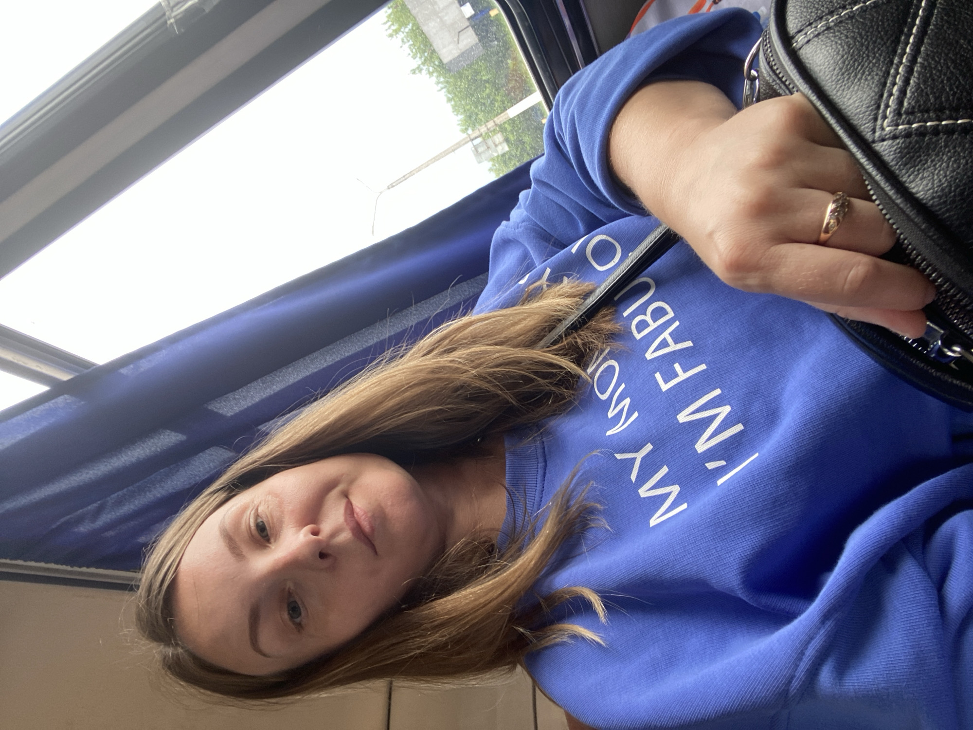 A woman with light brown hair is taking a selfie while sitting on a bus in Chaikovsky, Russia. She is wearing a blue sweatshirt with white text that reads "MY MOM I'M FABULOUS". She is holding a black bag. The background shows a window with blue curtains and trees and buildings visible outside.