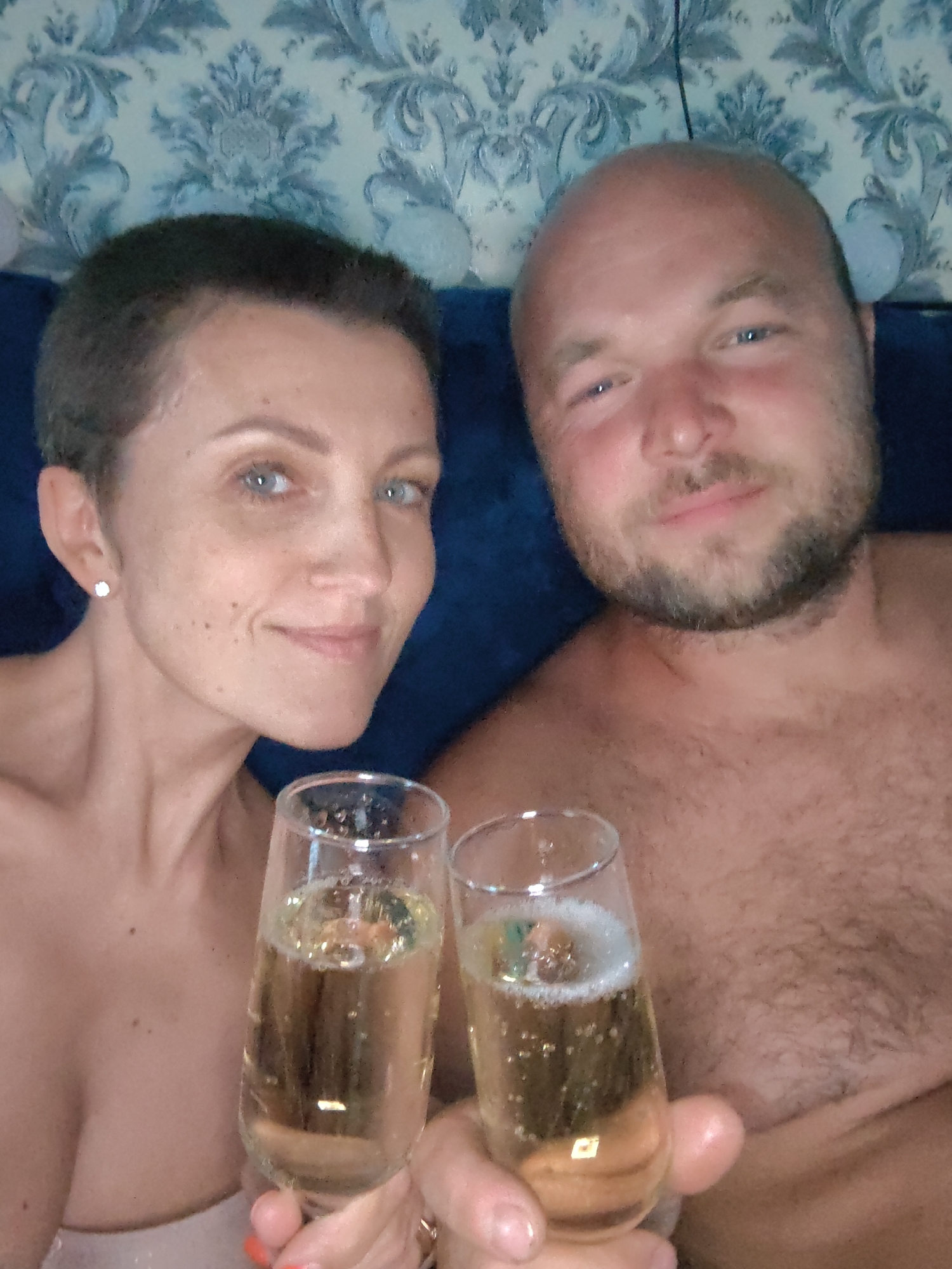 This is a close-up, overhead, selfie-style photograph of a man and a woman in what appears to be a domestic setting in Fanipal, Belarus. The man, with a shaved head and a beard, is on the right side of the frame, with his chest and face visible. The woman, with short dark hair and blue eyes, is on the left side of the frame, with her face and upper chest visible. Both are holding champagne flutes filled with a bubbly, golden liquid. The woman is wearing a small, sparkling earring. The background features a dark blue upholstered surface and a patterned wallpaper with blue and white motifs. The lighting is soft, suggesting an indoor environment, possibly a bedroom or living area. The overall tone of the image is intimate and celebratory.