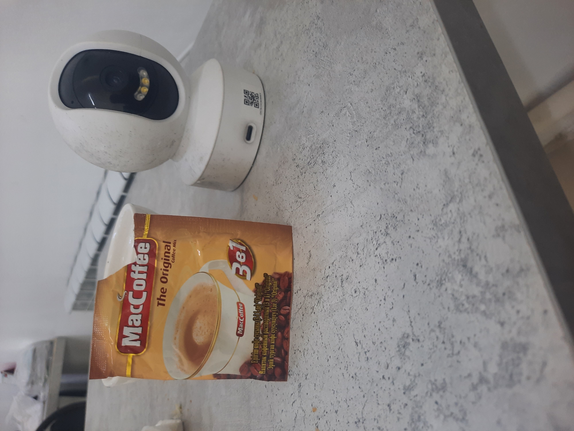 In Zhanaozen, Kazakhstan, a white security camera with a black lens sits on a textured gray countertop. To its left, a sachet of "MacCoffee The Original Coffee Mix" is partially visible. The sachet is gold and brown and depicts a cup of coffee and coffee beans. The background includes a white wall and a radiator.