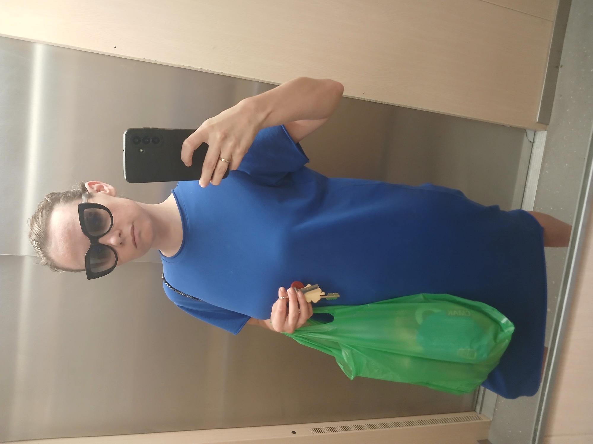 In an elevator with stainless steel walls and a wooden panel, a woman is taking a selfie. She is wearing large, black, cat-eye sunglasses and a bright blue dress. On her left arm, she is carrying a bright green plastic shopping bag. Her right hand, which is holding her phone, has a ring on her ring finger. She is also holding a set of keys in her left hand, which is near the shopping bag. The elevator floor appears to be tiled with a speckled pattern.