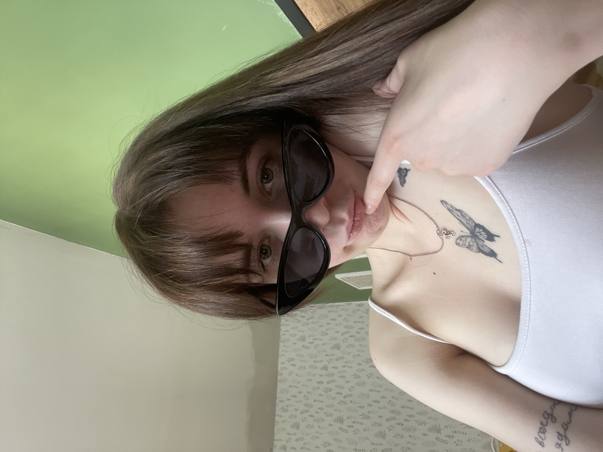 A young woman with brown hair and sunglasses is shown in a selfie. She is wearing a white tank top and has tattoos of butterflies on her chest and left arm. The background consists of a green wall and a floral-patterned wall or curtain. The location is likely indoors, but there are no further details available to determine if it is in the city of Chaikovsky, Russia.