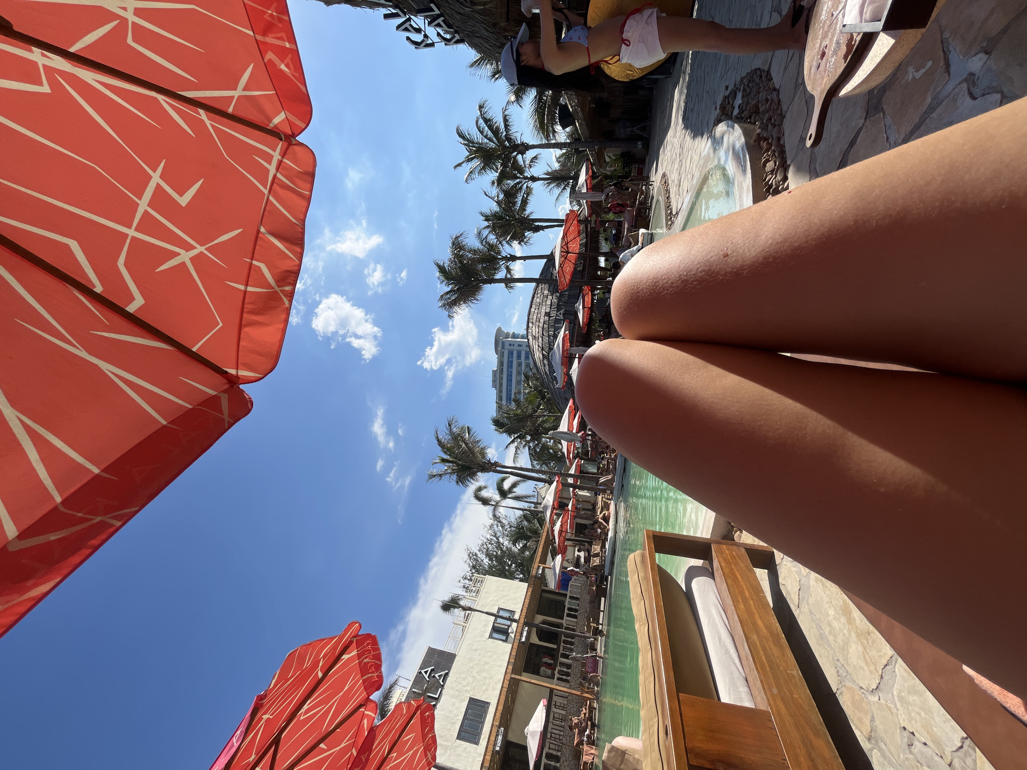 The image captures a vibrant and sunny poolside scene, likely at a resort in Đà Nẵng, Vietnam, viewed from a first-person perspective. In the immediate foreground, the tanned legs of a person are prominently displayed, suggesting they are relaxing on a lounge chair. A large, bright red umbrella with white abstract patterns covers the upper left portion of the frame, providing shade, with another similar umbrella partially visible at the bottom left.
The setting features an inviting swimming pool with clear turquoise water, surrounded by tropical elements. Several tall palm trees punctuate the mid-ground, enhancing the vacation ambiance. Beyond the pool area, various buildings are visible, including one with partially visible white text that appears to read "AALI" or "AHLI". Other individuals can be seen in the background, some seated at tables or under umbrellas, contributing to the leisurely atmosphere. In the upper right corner, another person is partially visible, seated at a table with a wooden serving board, possibly holding drinks or snacks. The sky is a brilliant blue with scattered white clouds, indicating a clear, bright, and sunny day, suggesting mid-morning or early afternoon due to the strong light. The overall mood is one of relaxation and enjoyment at a tropical destination.