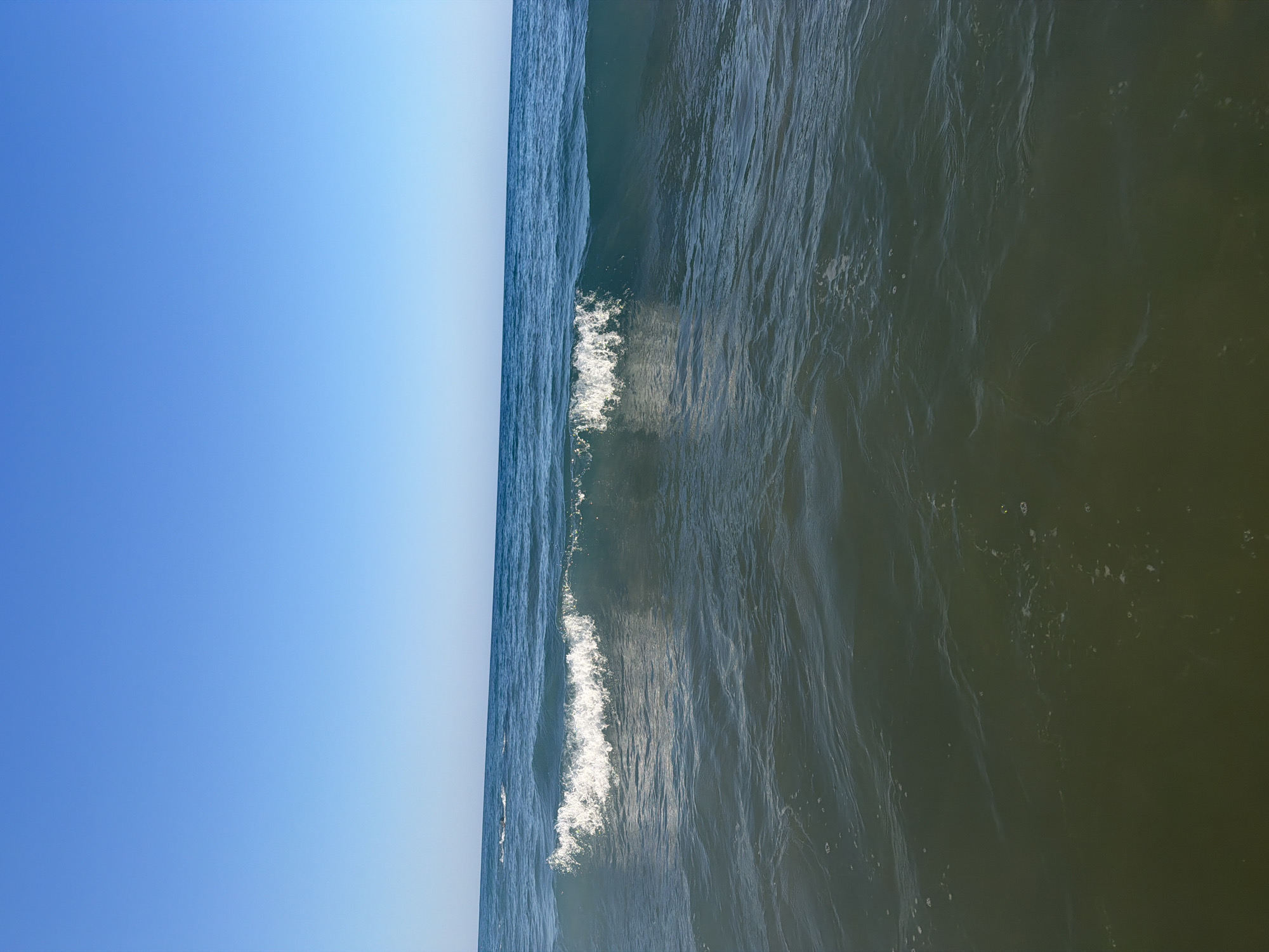 This is a photograph taken from a low vantage point, appearing to be within the water itself, looking out towards the sea. The image is dominated by the expanse of the clear blue sky, which occupies the top half of the frame. Below the sky, the deep blue-green ocean stretches across the horizon, with a gentle wave breaking in the middle ground. The wave is captured in motion, with its crest frothing into white foam. The water in the foreground is a darker, murkier green, showing subtle ripples and the occasional small bubble. The overall impression is one of a calm, clear day at the beach. The location is identified as Georgioupoli, Greece.