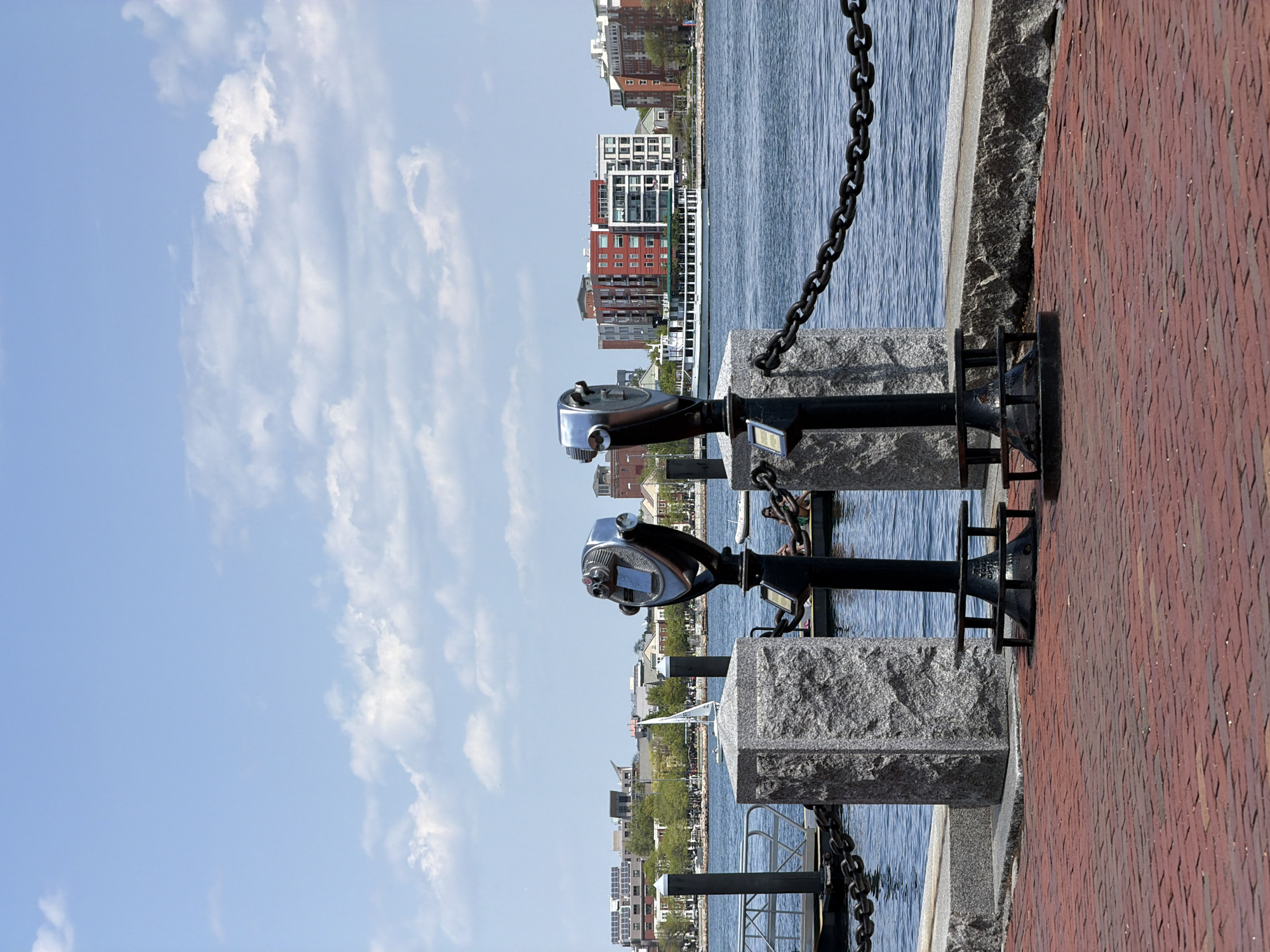 This eye-level shot captures two coin-operated binocular telescopes on a waterfront promenade, with the skyline of Boston, Massachusetts, in the background. The telescopes are mounted on black metal stands fixed to granite blocks, which are placed along a brick pathway. A heavy, black chain runs parallel to the granite blocks, acting as a safety barrier between the promenade and the water.
The sky is light blue with scattered white clouds. Across the water, various buildings compose the Boston skyline, including residential apartments and commercial structures. The water reflects the colors of the sky and buildings, creating a rippled effect. The image is taken during daylight, as indicated by the clear sky and natural lighting.