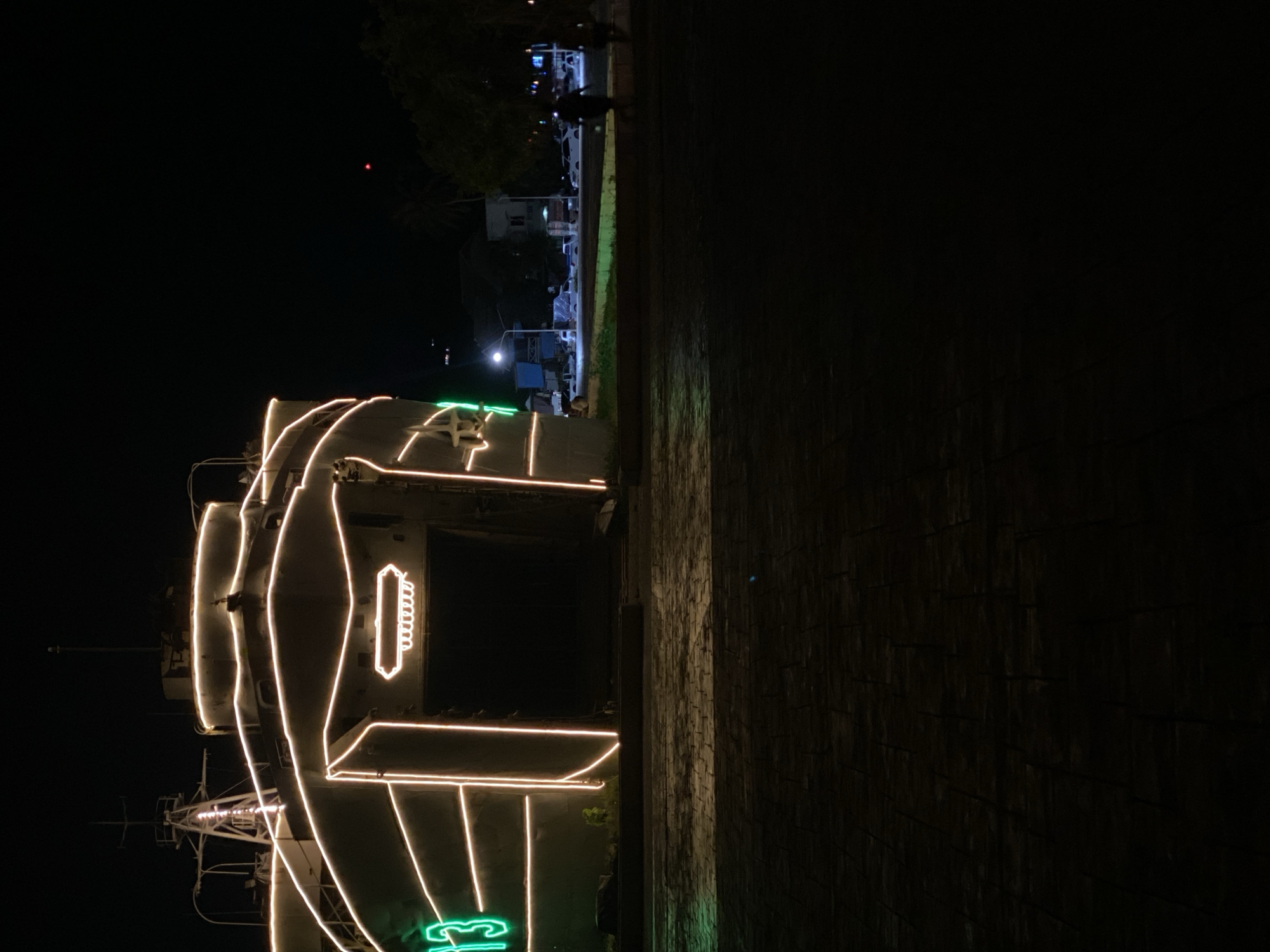 The image shows a dark night scene in Baan Don Sai, Thailand. The USS Selma Montgomery is decorated with neon lights. A paved area is on the right. In the background, there are trees, buildings, and streetlights. There are two people walking in the upper part of the image.