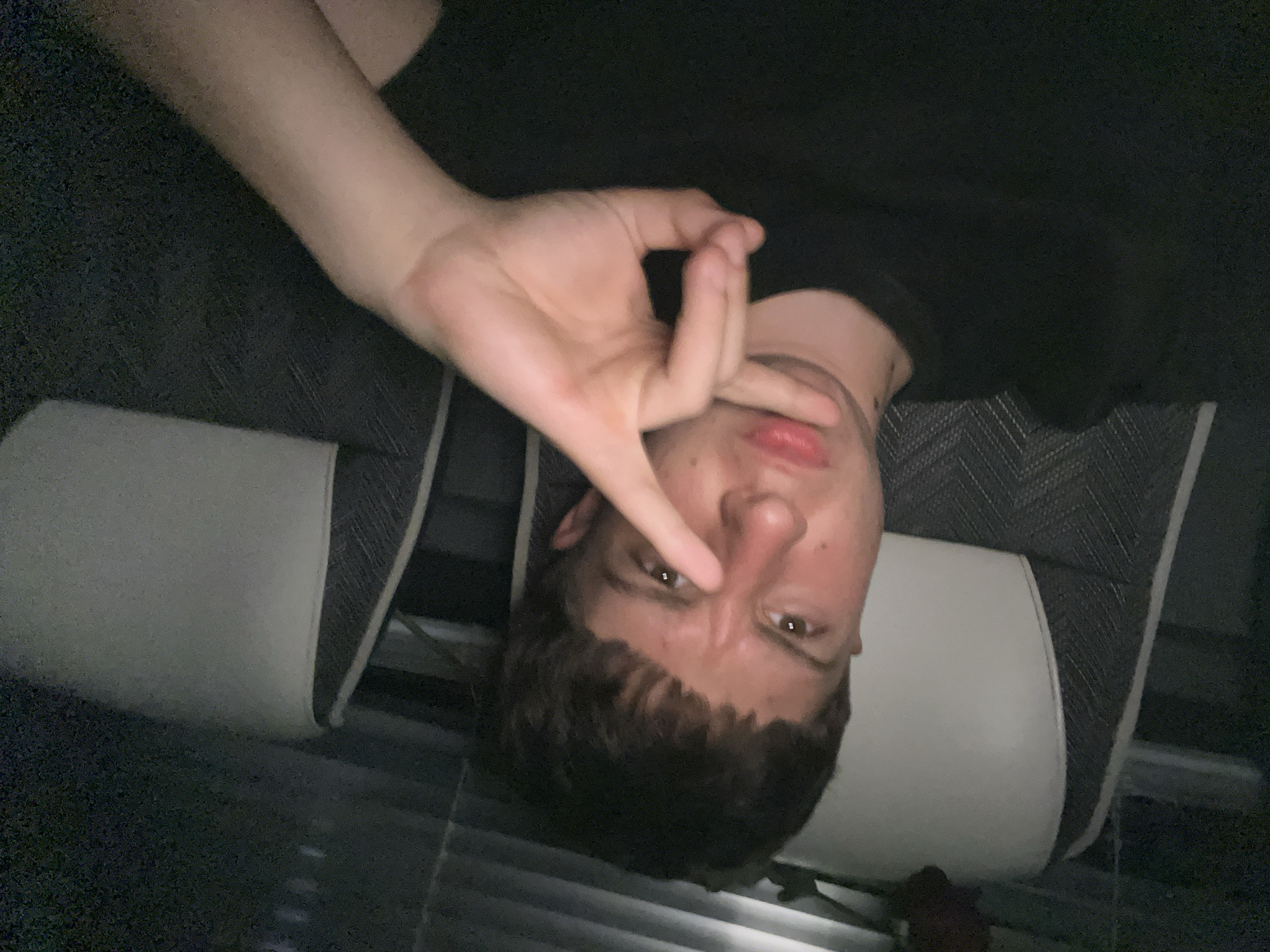 A person with brown hair, light skin, and brown eyes is lying upside down, making an "okay" hand gesture over their left eye. The person is wearing a dark-colored shirt or top. The background appears to be furniture, including a seat with white cushions and dark gray upholstery. The location is unknown and there is no evidence to suggest that it is in Saraia, Bulgaria.