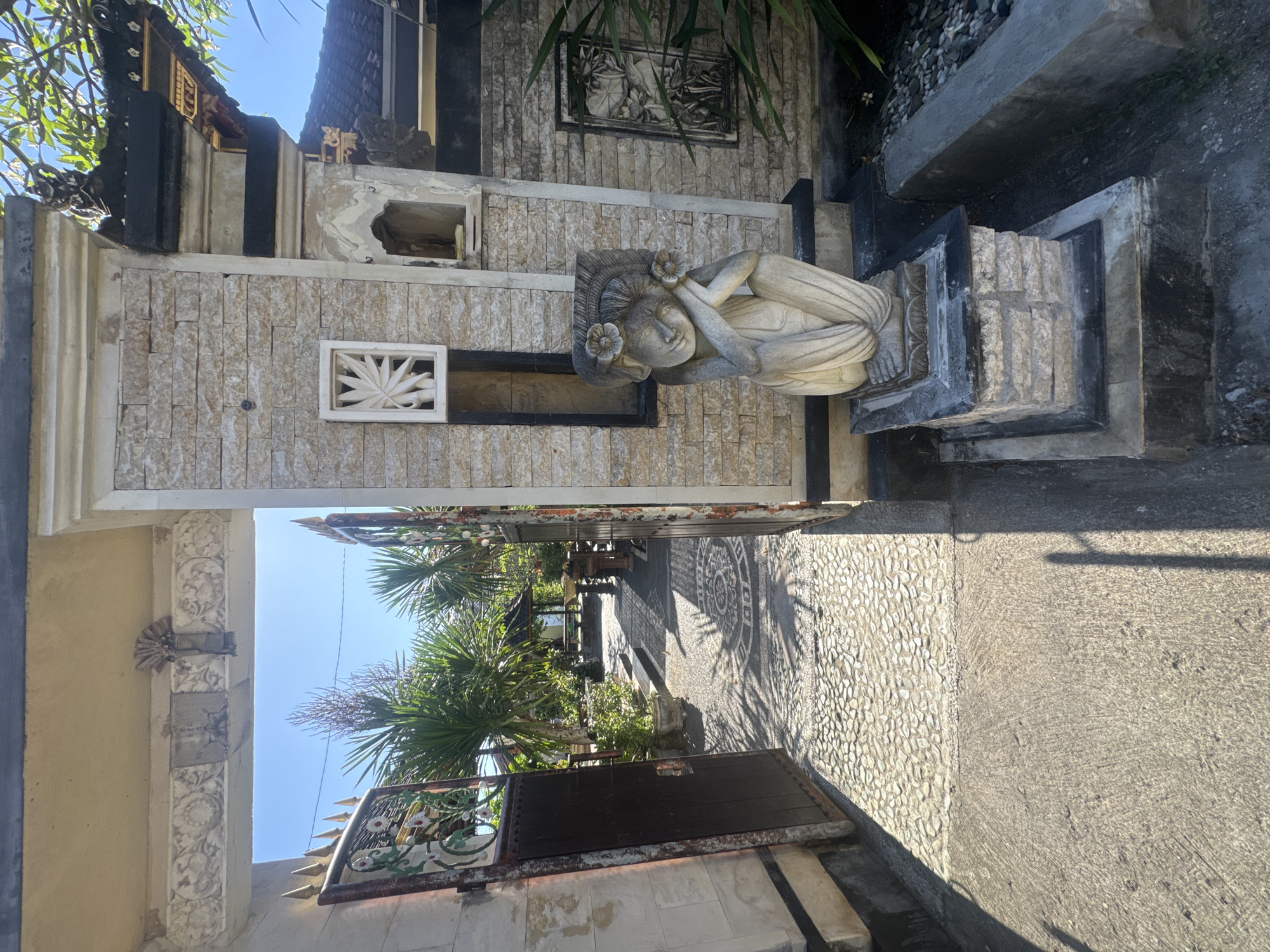 The image depicts a street scene in Kuta, Indonesia, featuring Balinese architecture and decor. A stone wall adorned with a sleeping woman statue and a decorative window runs along the edge of a pebbled walkway. An open gate leads to a garden area with palm trees and other greenery. The architecture incorporates intricate carvings and stonework, typical of Balinese style. Sunlight casts shadows across the walkway. No people are visible.