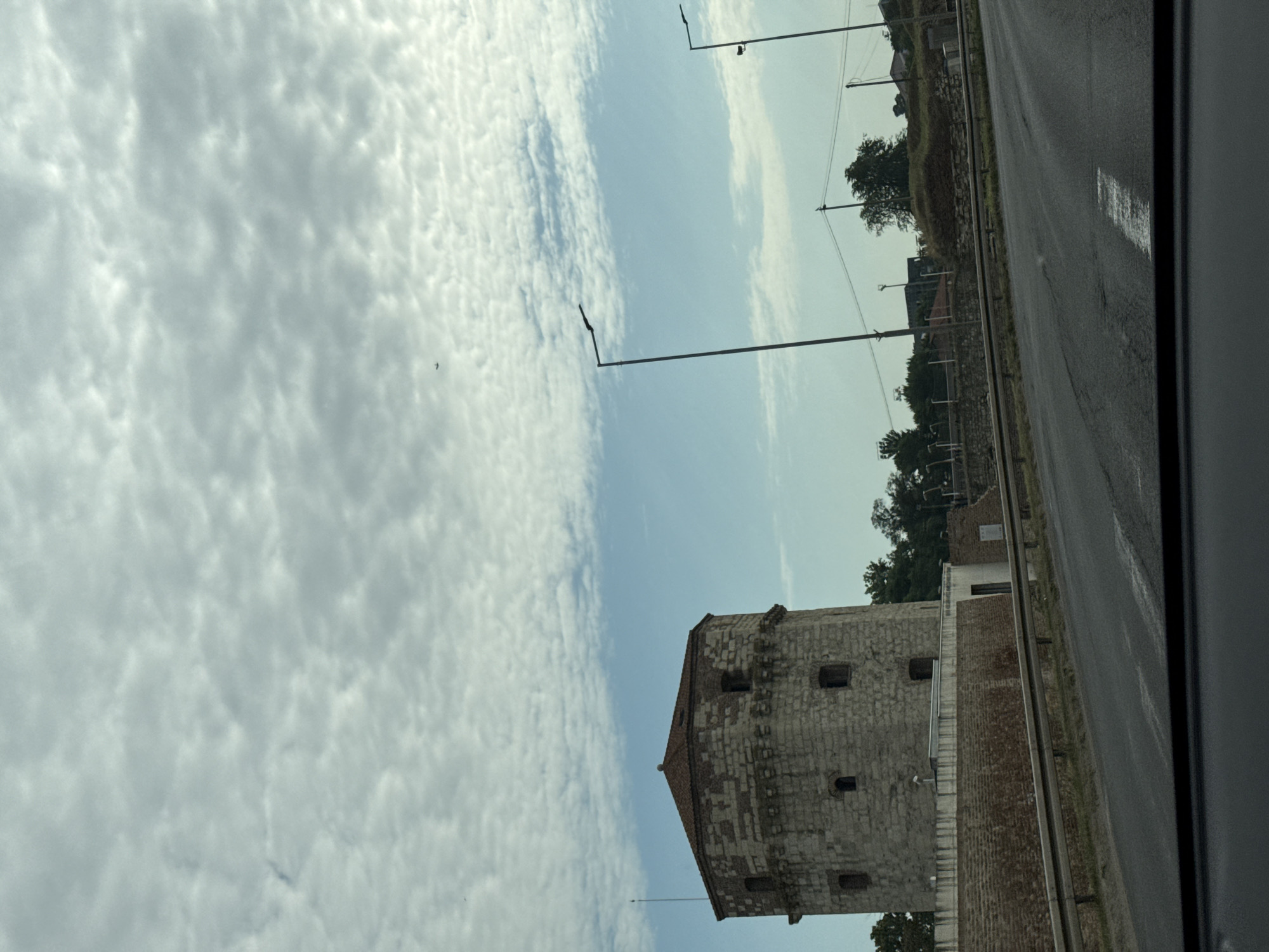 The image shows a view from a car driving on a road in Belgrade, Serbia. In the background, the prominent subject is the white stone tower of the Belgrade Fortress, also known as Kalemegdan Fortress. The tower is octagonal in shape with a reddish-brown tiled roof and several small, arched windows. The tower is surrounded by a lower brick wall.
The sky above is partially cloudy with patches of blue. Two tall, modern streetlights with dark fixtures are visible on the right side of the road. Power lines and poles are also present in the middle ground. Trees and some buildings are scattered along the horizon behind the fortress and the road. The road itself is asphalt and appears to be a multi-lane highway, with a metal guardrail separating it from the area with the fortress. The overall scene depicts an urban landscape with a historic structure juxtaposed with modern infrastructure.