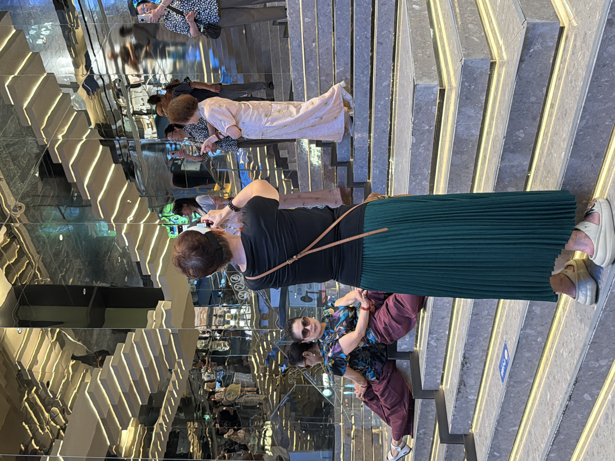 This image depicts a group of people on a grand, mirrored staircase in a modern building, likely a shopping mall or hotel, in Yuzhong District, China. The staircase itself is a prominent feature, with multiple levels and illuminated edges. The mirrored walls reflect the architecture and the people, creating a sense of depth and visual complexity.
In the foreground, a woman with short, dark hair, wearing a black t-shirt and a long, dark green pleated skirt, is standing and holding a smartphone, appearing to take a photo. To her left, two women are seated on a lower step of the staircase. The woman in the front is wearing a black, colorful patterned top and dark maroon pants, with large sunglasses and her arms resting on her knees. Beside her, another woman, with her back mostly to the camera, wears a similar outfit.
Further up the stairs, several other individuals are visible, some interacting and taking pictures with their phones. One woman in a light pink, long dress is standing and looking around. The overall atmosphere suggests a tourist or social gathering, with a focus on capturing the experience through photography. The mirrored surfaces and geometric design of the staircase indicate a contemporary architectural style, common in urban commercial spaces. The lighting appears to be a mix of ambient interior light and the illuminated staircase itself, creating bright highlights and reflective surfaces.