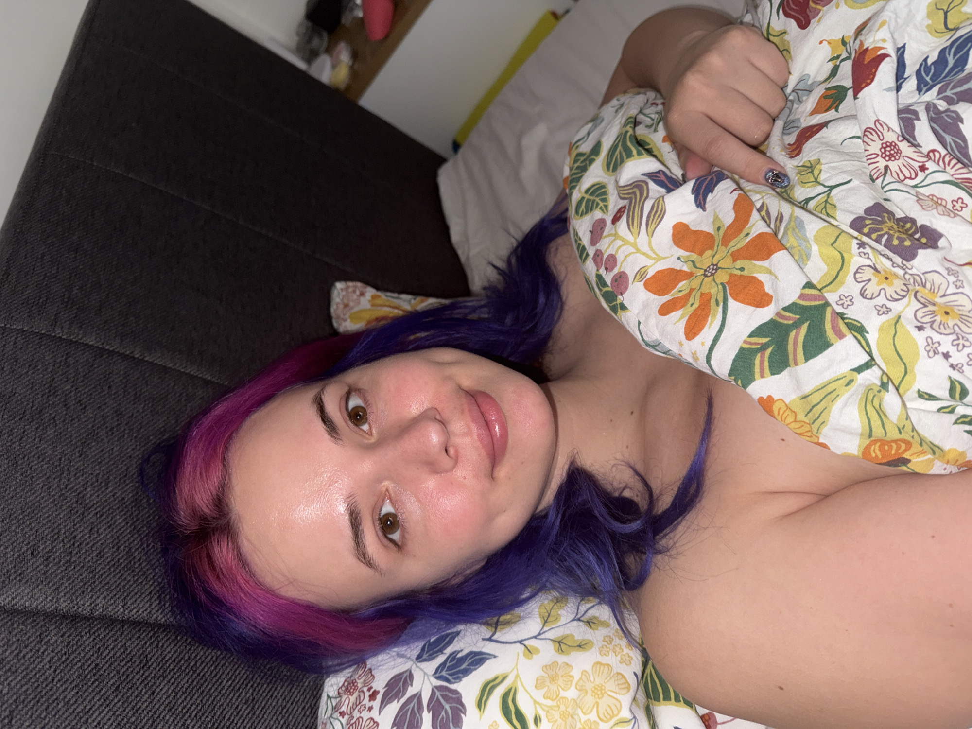 A person with vibrant, two-toned hair — purple transitioning to pink/magenta at the roots — is captured in a close-up, eye-level shot while lying in a bed. The individual looks directly at the camera with a relaxed and soft expression, a subtle smile playing on their lips. Their fair skin shows a noticeable sheen on the forehead.
The setting appears to be a bedroom, likely in the City of Zagreb, Croatia. The person is partially covered by a duvet and resting their head on a pillow, both featuring a lively floral pattern with yellow, orange, green, red, and purple elements against a white background. One hand is visible, gently gripping the duvet, and a fingernail on the visible hand showcases a distinct blue and silver glitter design.
To the left, a dark grey, textured fabric headboard is visible, indicating the bed's structure. The background is softly lit, suggesting evening or nighttime, with a light-colored wall and a shelf holding various, indistinct items. The overall atmosphere conveys comfort and ease. There is no discernible text within the image.