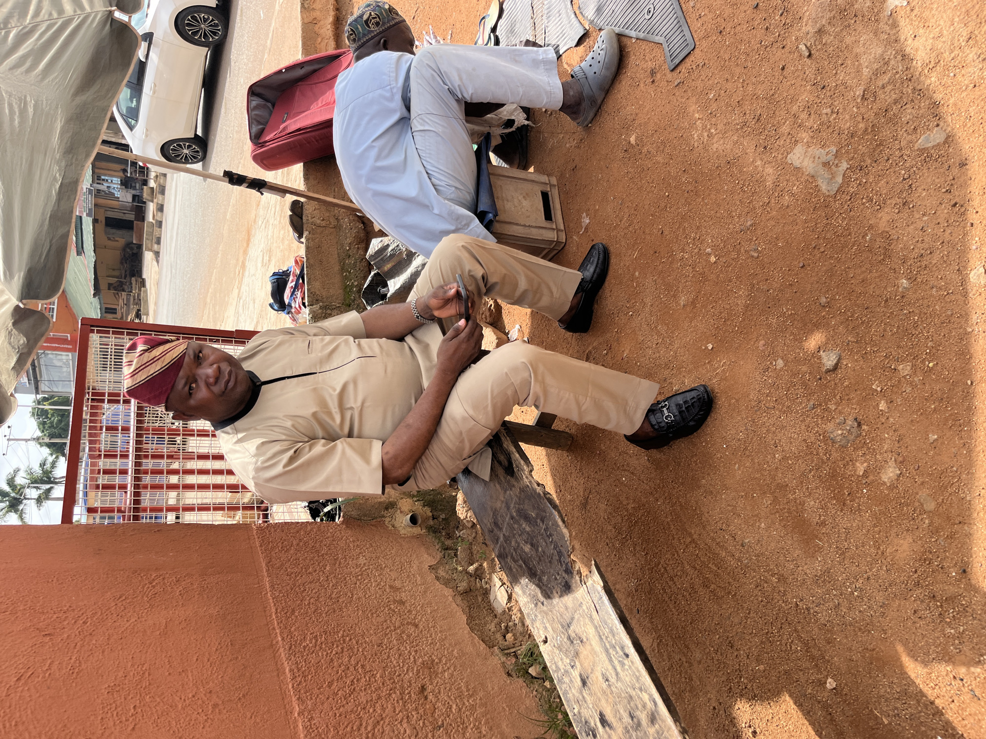 In the city of Bukuru, Nigeria, an adult male wearing a tan traditional outfit and a patterned red and gold cap sits on a rustic wooden bench on a dirt surface. He holds a dark-colored smartphone in his left hand. To his right, another adult male, also wearing a light blue traditional outfit and a patterned cap, is crouched down, tending to something on the ground. A red suitcase is positioned to the right of the crouching man, and a wooden crate is partially visible underneath him. A light-colored umbrella provides shade over the scene. In the background, a white car is parked on a paved road, and buildings with red and white fencing are visible. The environment is dusty, with scattered stones and sparse vegetation.