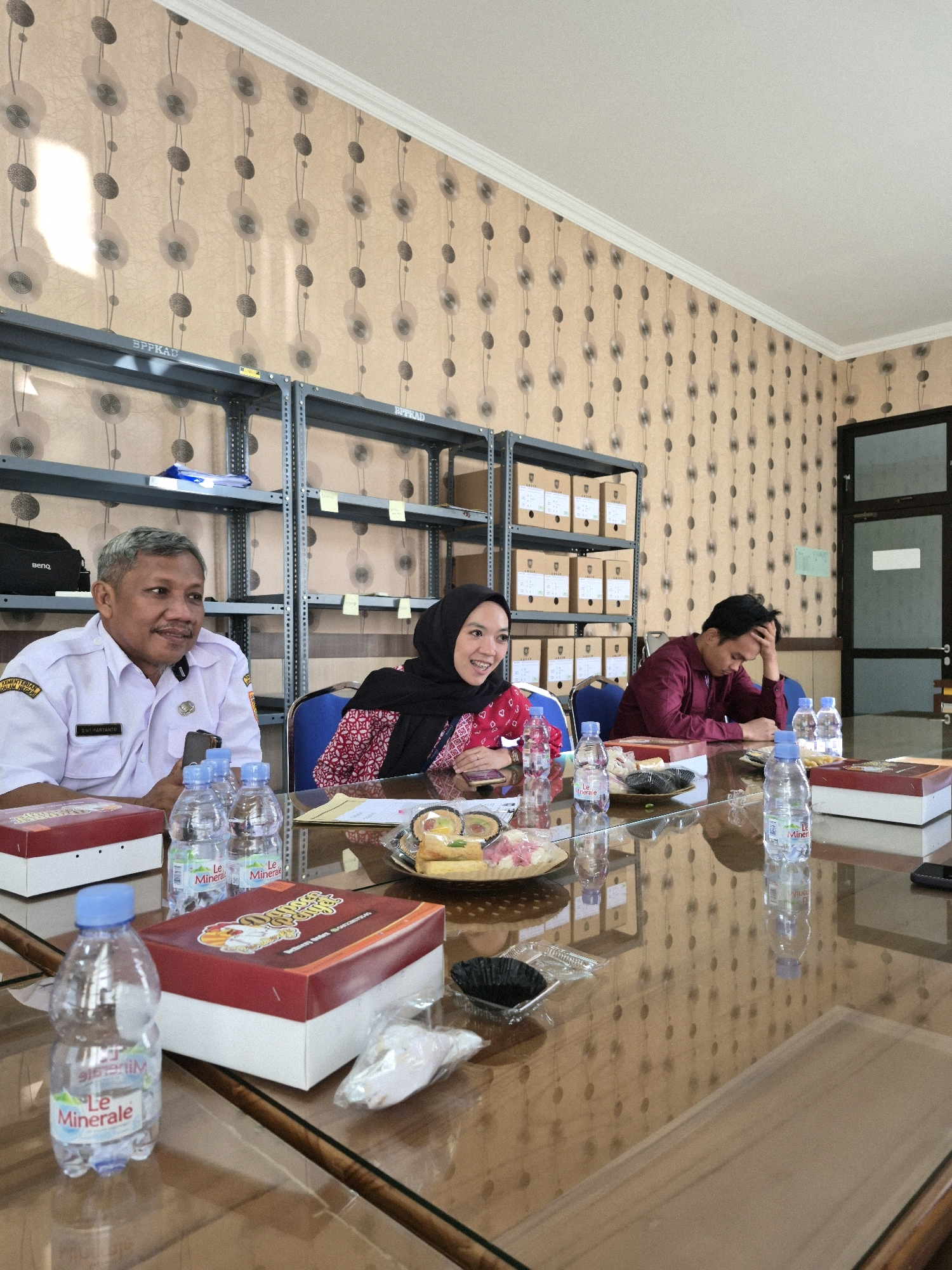 The image depicts an indoor scene, likely a meeting room or office, in Purworejo, Indonesia, given the context provided. Three individuals are seated at a polished wooden table with a glass surface, reflecting the patterned wallpaper and ceiling lights. The wallpaper features a repeating motif of stylized circular forms on a light beige background. Metal shelving units filled with boxes are visible in the background.
In the foreground and on the table, several bottled waters, some with "Le Minerale" labels, are placed. There are also food boxes, one red and white with a graphic design, and a small plate with snacks.
On the left, a man wearing a white uniform shirt with a name tag that reads "Eri Hartanto" and an emblem on the chest is seated. He has graying hair and is looking to his right, holding a dark-colored phone.
In the middle, a woman wearing a black hijab and a patterned red and white top is smiling and looking towards the right. She is also holding a mobile phone.
On the right, a man in a maroon shirt is seated with his head resting on his hand, appearing to be either in thought or perhaps tired.
The overall atmosphere suggests a casual business meeting or a gathering. The lighting appears to be a mix of overhead fluorescent lights and natural light coming from a window or opening on the left, casting a bright rectangular patch on the wallpaper.