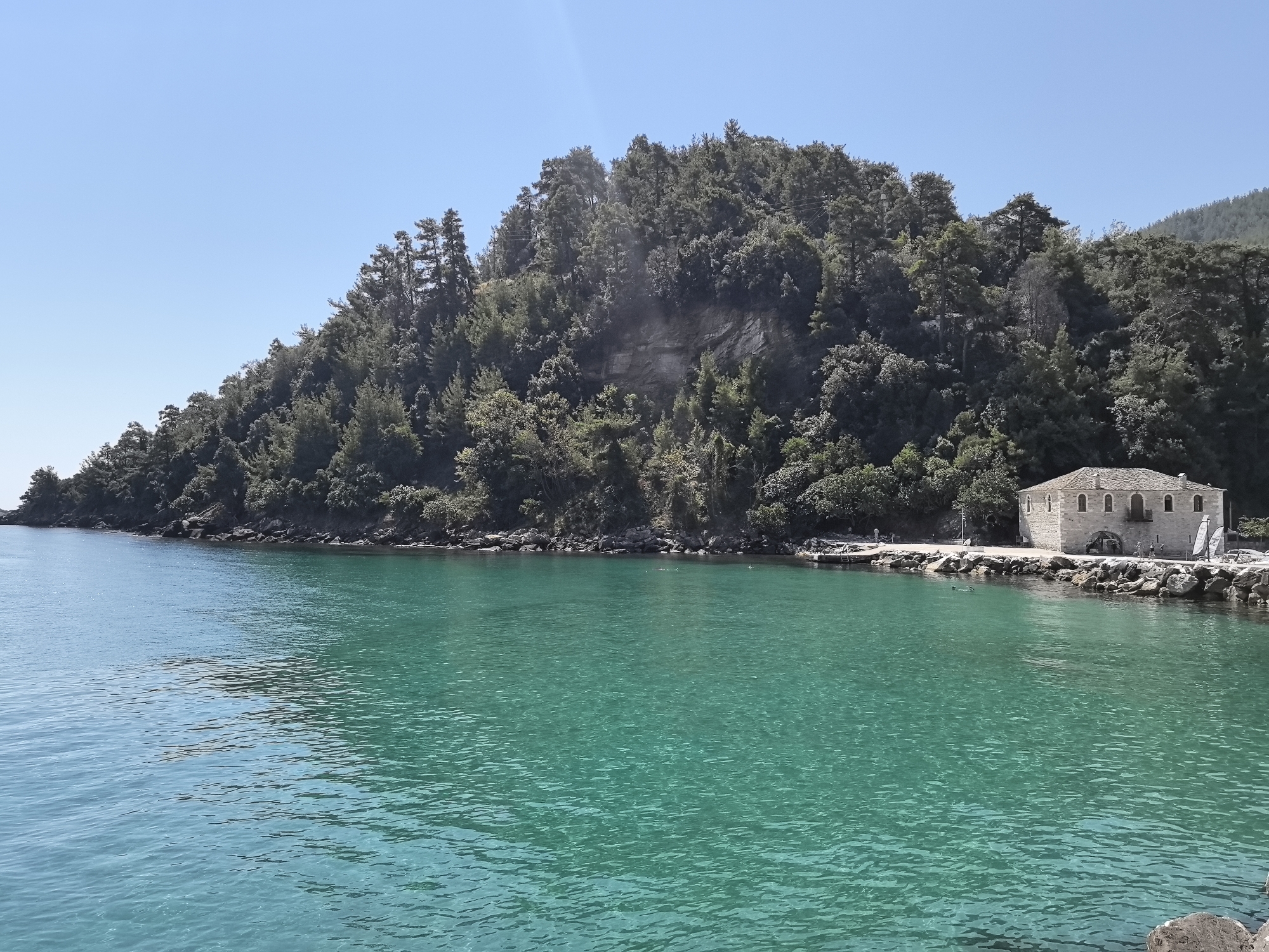 A clear, turquoise body of water dominates the foreground of the image. The water is calm with gentle ripples reflecting the bright sunlight. In the background, a lush, tree-covered hillside slopes down to the coastline. Along the shore, a stone building with arched doorways and numerous windows stands prominently. Several people are visible on the shore near the building and some are in the water, appearing to be swimming or snorkeling. A rocky coastline borders the water, with a few small boats docked near the building. The sky is a clear, cloudless blue. This scene is located in Skala Potamias, Greece.
