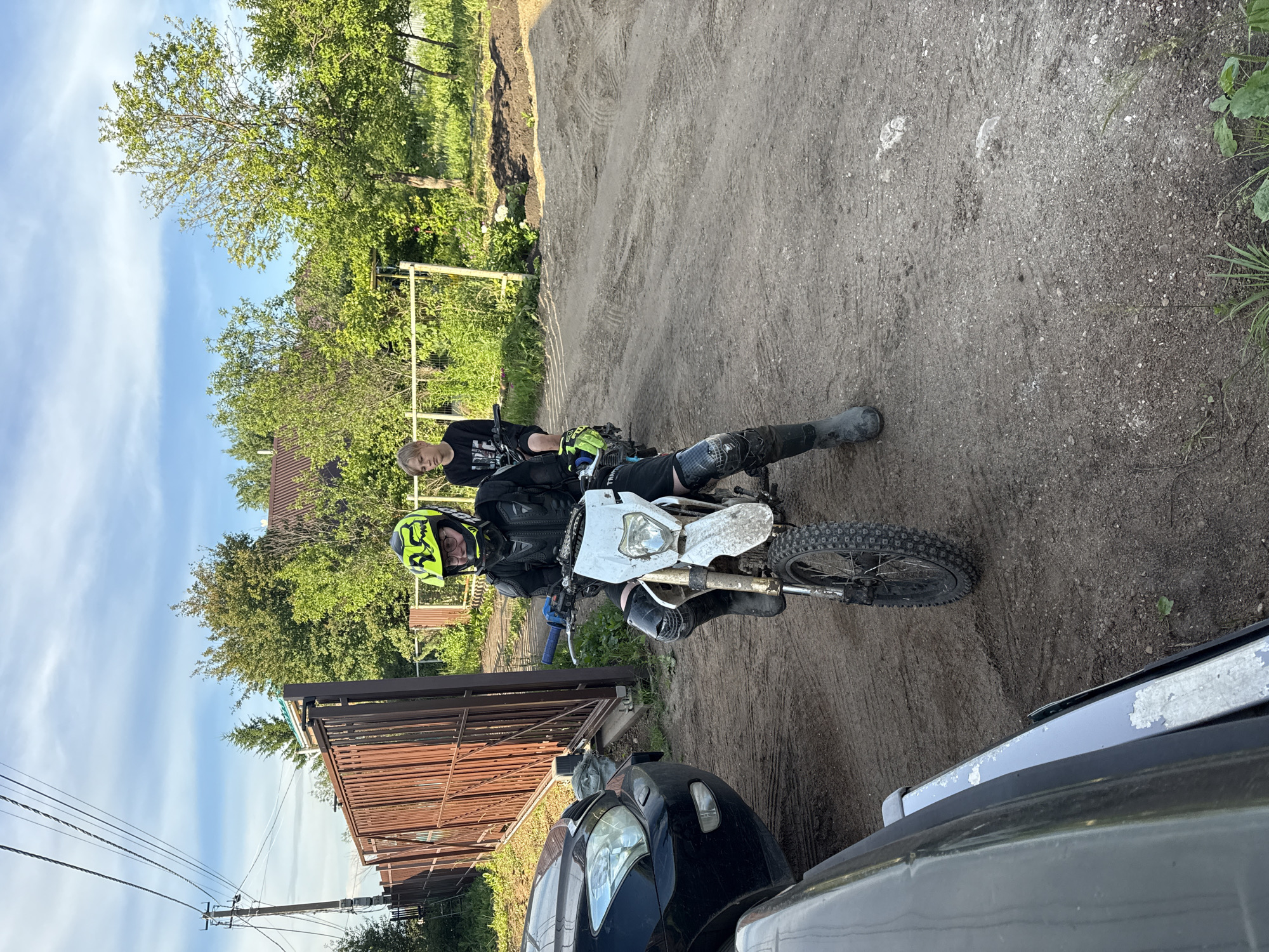 Two young males are on a dirt track, with one sitting on a dirt bike and the other standing behind. The individual on the dirt bike is wearing a yellow and black helmet, black racing gear, and knee pads. The second individual is wearing a black long-sleeved shirt. A black car is parked to the left of the scene, and a brown metal gate is visible in the background. The environment appears to be a rural area with trees and vegetation. The ground is dirt and dusty. The sky is blue with some clouds.