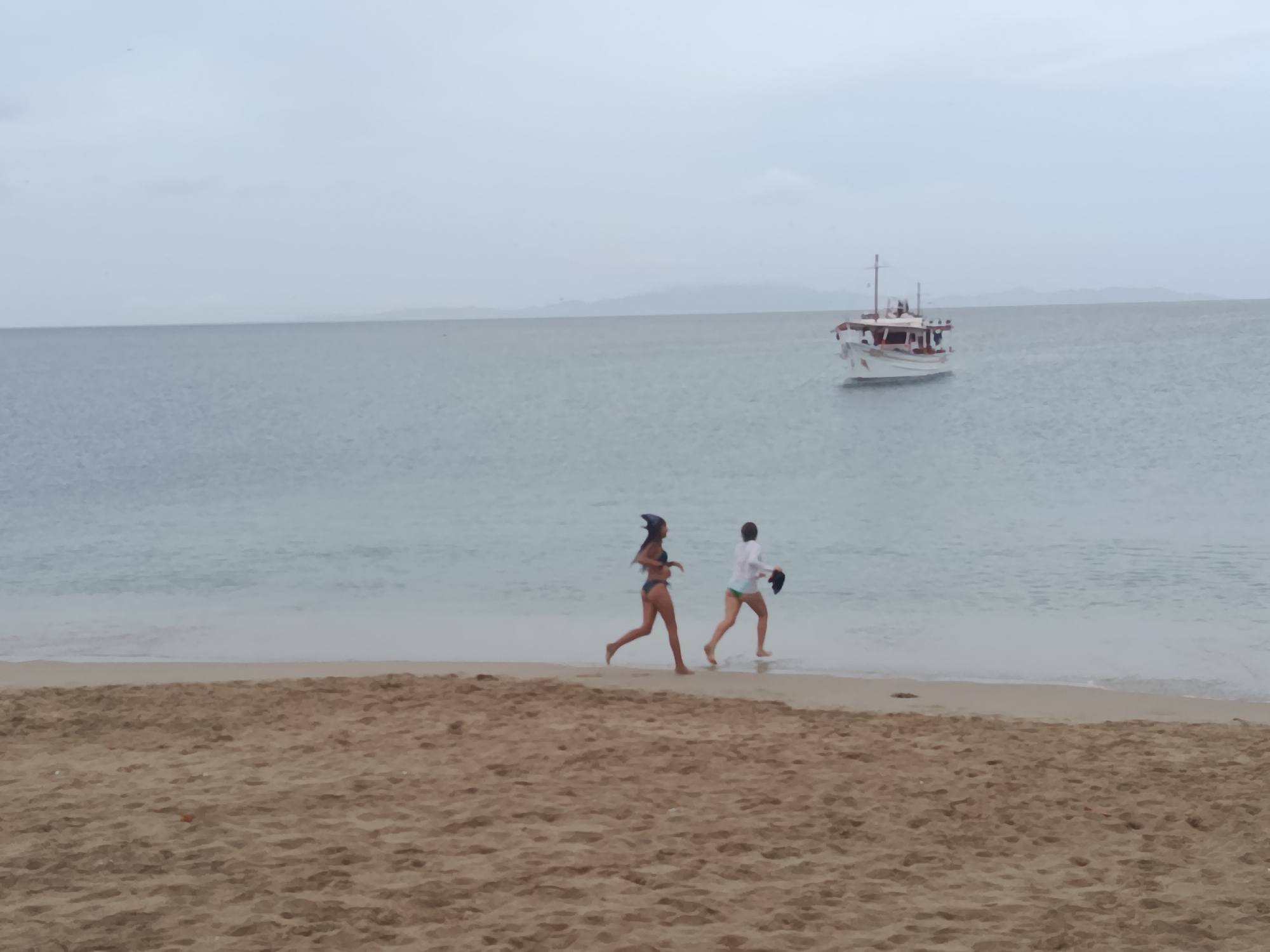 Two individuals are running along a sandy beach near the water's edge in Juan Griego, Venezuela. The person in front, on the left, is wearing a dark bikini and a dark headscarf, running with their right leg forward. The person behind, on the right, is wearing a white shirt and green bottoms, also running with their right leg forward, holding dark items in their right hand. The sea is calm and gray, with a light blue hue, and a white boat with red accents is visible in the distance on the water. The sky is overcast and gray. The sand in the foreground is textured and appears dry.