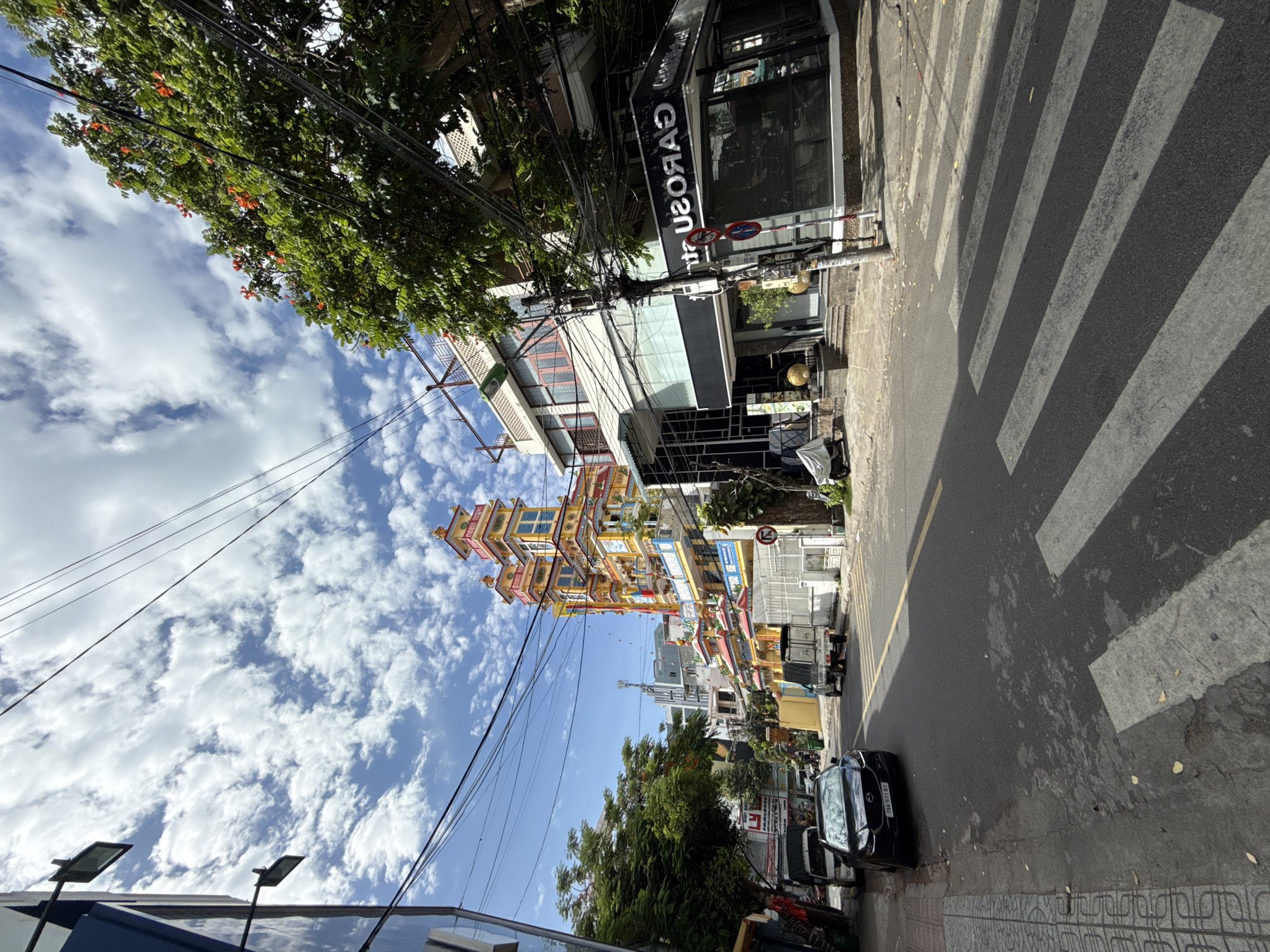 The image depicts a street scene in Nha Trang, Vietnam. A colorful, multi-tiered Buddhist temple with yellow and red accents dominates the right side of the frame. In front of the temple, a narrow street runs towards the viewer, with a zebra crossing in the foreground. Several vehicles are parked along the left side of the street, including a dark-colored sedan and a white van. Buildings line both sides of the street, with a shop featuring a black and white sign that reads "GAROOS" visible on the right. Lush green trees with orange flowers are present on the right side of the street, and a tangle of electrical wires crisscrosses the sky. The sky is blue with scattered white clouds. The ground is asphalt on the street and paved with tiles on the sidewalk. The overall impression is one of a vibrant urban environment with a strong cultural presence.