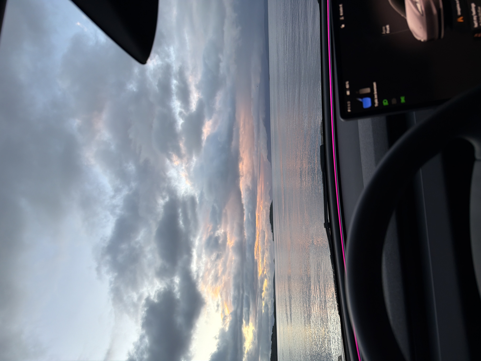 The image shows a view from inside a vehicle looking out at a body of water and a cloudy sky at sunset. The water reflects the colors of the sky, which range from pale blue to orange and pink. There are islands and mountains visible in the distance. The dashboard of the car is visible in the foreground, with a steering wheel and a touchscreen display. A pink light strip runs along the dashboard. The scene appears to be located near the city of Harstad, Norway, as suggested by the presence of islands and mountains characteristic of the Norwegian coast. The overall impression is one of a serene and beautiful natural landscape.