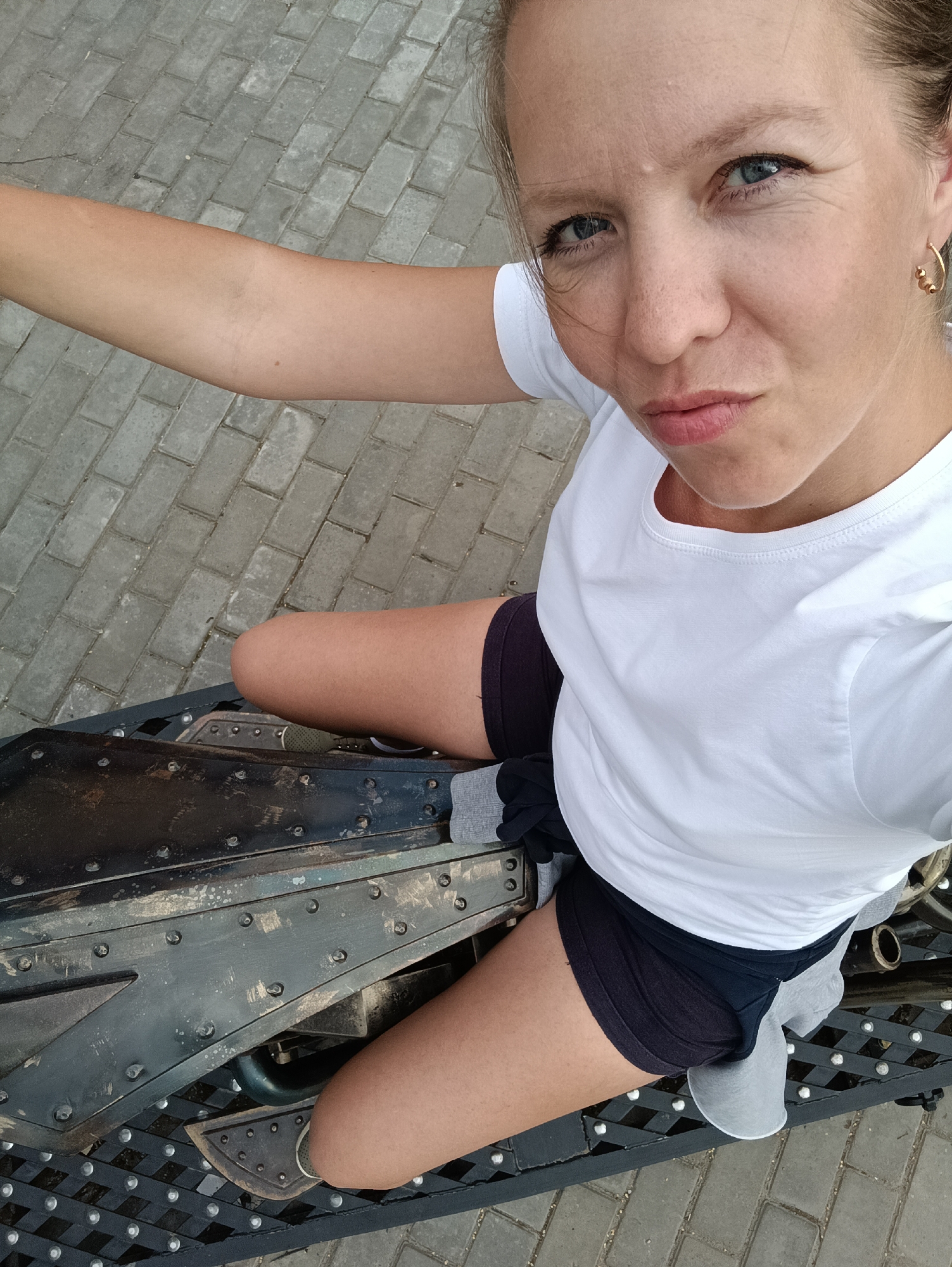 A woman is captured in a high-angle selfie, her body positioned on what appears to be part of a historical or artistic installation. She is wearing a white t-shirt and dark shorts. Her right arm is extended, holding the camera. The background consists of a gray paved walkway. Her facial expression is a playful pout. The installation is metallic and appears weathered, with visible rivets and some dirt or grime. The location is identified as Chaikovsky, Russia.