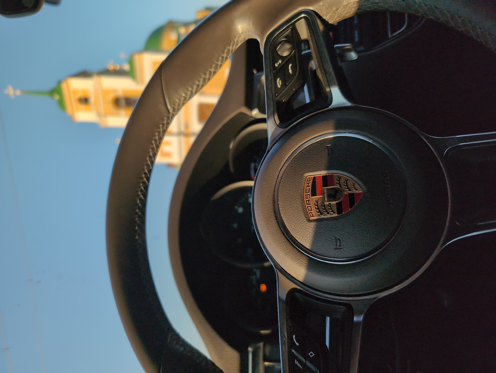 The image shows the steering wheel of a Porsche with the Porsche logo prominently displayed in the center. Through the windshield, a blurred building with yellow and green domes is visible. The building's architecture suggests an Orthodox church. Based on the city specified, the church is likely located in Мазунино, Russia. The sky is visible above the building and appears blue.