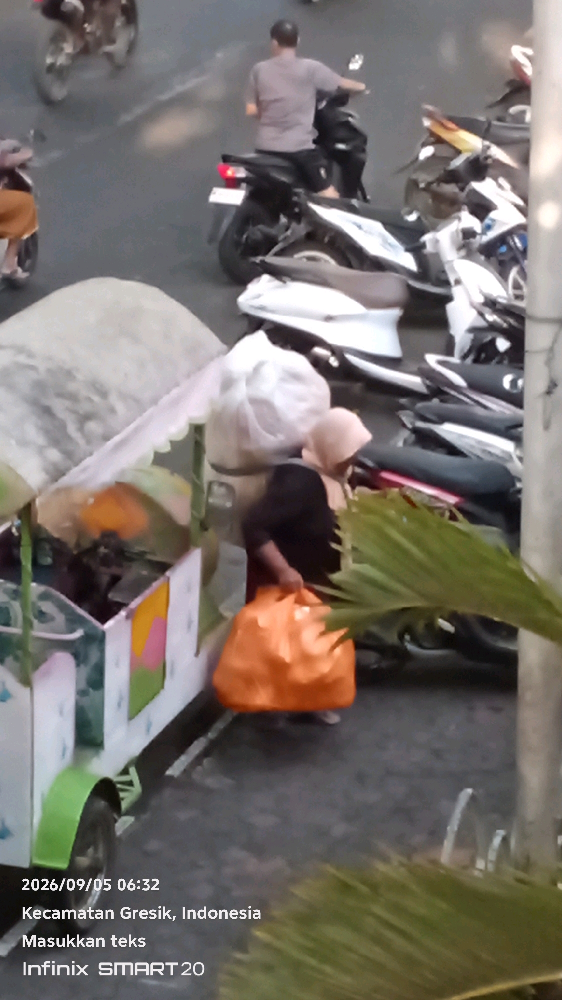 A woman wearing a light-colored hijab and a dark top, carrying an orange bag, stands near a street cart. The cart has a white canopy with a scalloped edge and appears to be decorated with colorful panels. In the background, several motorcycles are parked or in motion on a paved street. The scene is illuminated by natural daylight. A timestamp in the lower left corner reads "2026/09/05 06:32" and indicates the location as "Kecamatan Gresik, Indonesia" with the text "Masukkan teks" and "Infinix SMART 20".