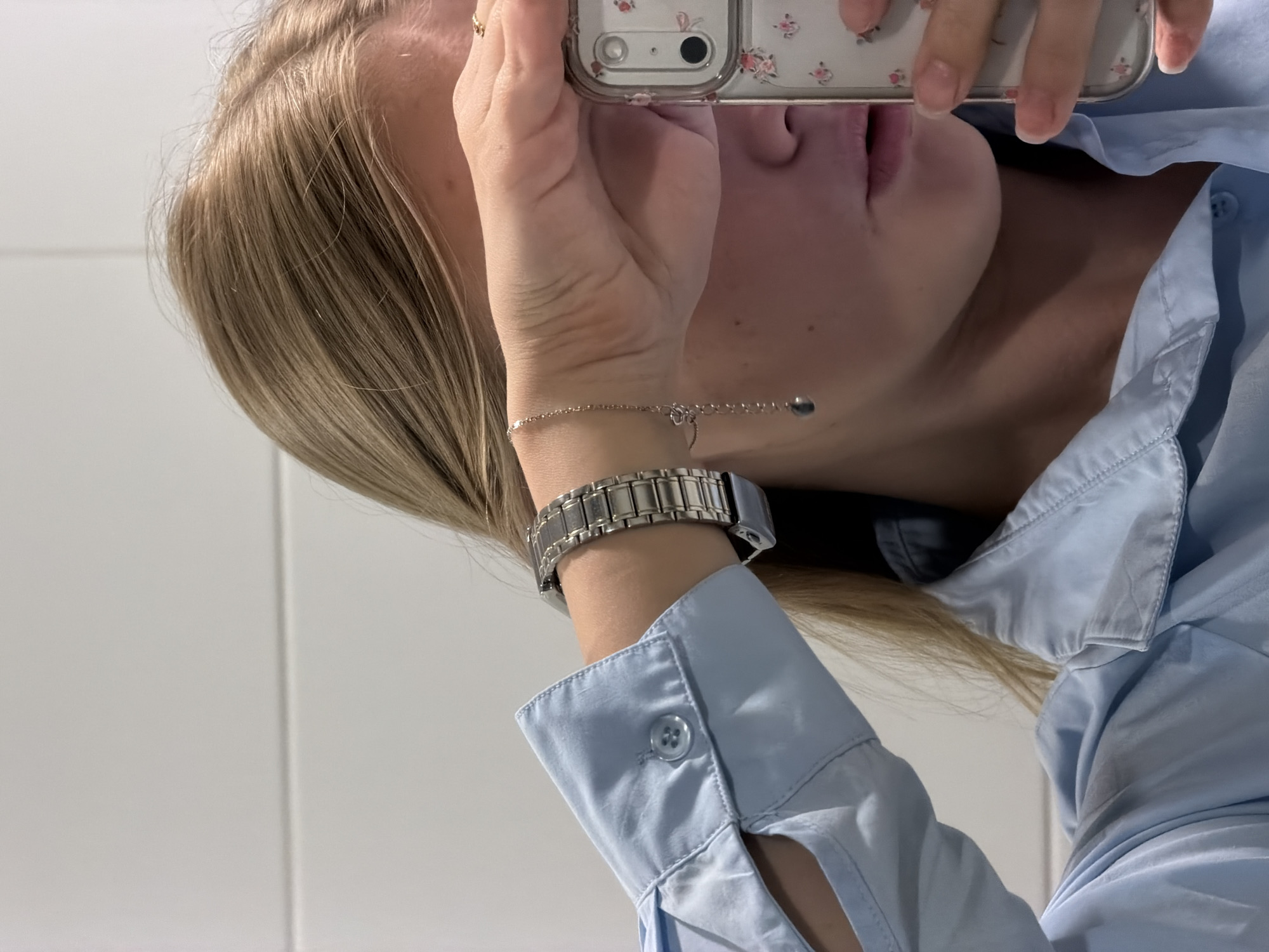 A person is holding a mobile phone up to take a selfie. The person has blonde hair and is wearing a light blue button-down shirt. The person is wearing a watch and a delicate bracelet on their left wrist. The background appears to be a white tiled wall. The location in the city of Izhevsk, Russia, cannot be determined from the image.