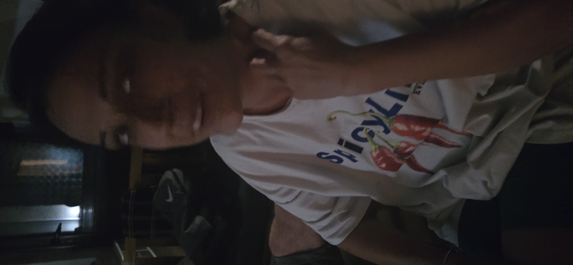 A person is captured in a close-up, indoor shot, likely in Govedare, Bulgaria, under low lighting conditions, suggesting evening or night. The individual, primarily visible from the chest up, has dark hair and is looking slightly upwards and to their left with a subtle smile. One hand is raised near their head, almost touching their ear. They are wearing a light-colored, possibly white, short-sleeved t-shirt featuring a graphic design. The visible text on the shirt includes the word "SpicyLi" in blue letters, alongside an illustration of two red chili peppers with green stems. The background is dark and out of focus, with hints of reflective surfaces or appliances, possibly in a kitchen setting, including a light source from what might be an oven or microwave in the lower-left. The overall atmosphere appears casual and relaxed.