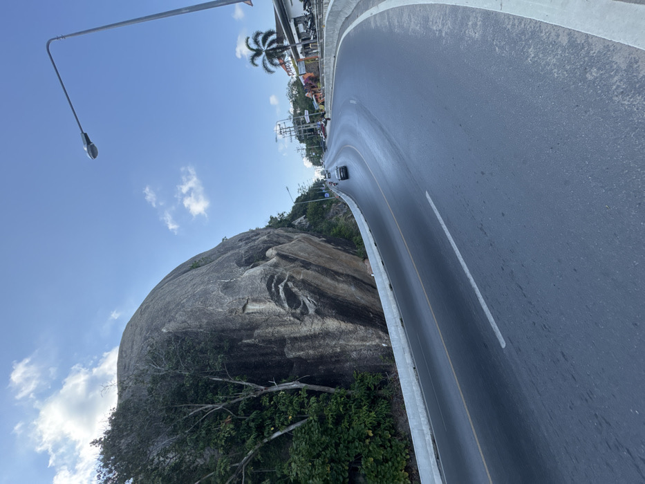 The image depicts a daytime scene in Baan Chaweng Noi, Thailand, featuring a curved two-lane asphalt road. On the left, a massive grey granite rock formation rises prominently, partially covered by dense green foliage and trees. The rock face exhibits vertical striations and a large, naturally formed, dark, eye-shaped depression. A light grey concrete barrier separates the road from the rock formation. The road, marked with a dashed white lane divider and a solid yellow line, curves to the right and ascends into the distance. Multiple vehicles, including a dark-colored sedan and several motorcycles, are observed on the road, proceeding away from the viewer. On the right side of the road, a tall grey metal street light pole extends upwards. Further down the right side, a series of commercial buildings with varied facades, some brightly colored, are visible behind another grey barrier. Palm trees and numerous utility poles with overhead power lines line this section of the roadside. The sky is clear blue with scattered white cumulus clouds.