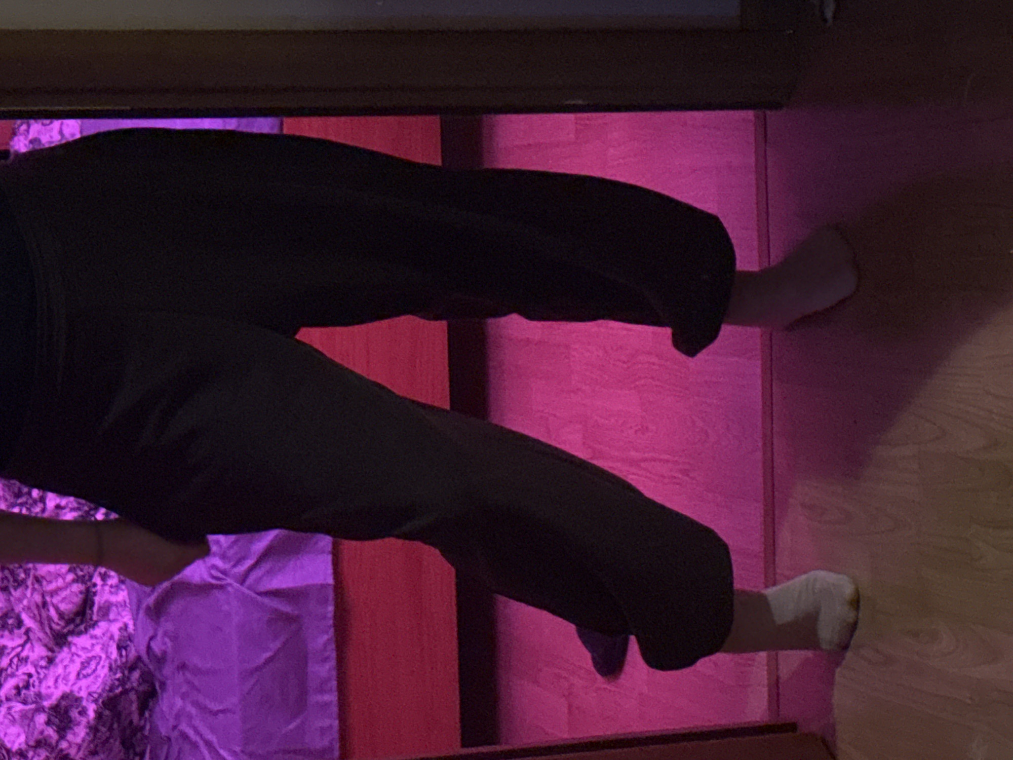 The image shows the lower body of a person wearing dark pants and a white sock on their left foot. The person is standing in what appears to be a bedroom with a bed covered in purple bedding and a pink light reflecting off the floor. The image does not contain any information about the location in the city of Moscow, Russia.