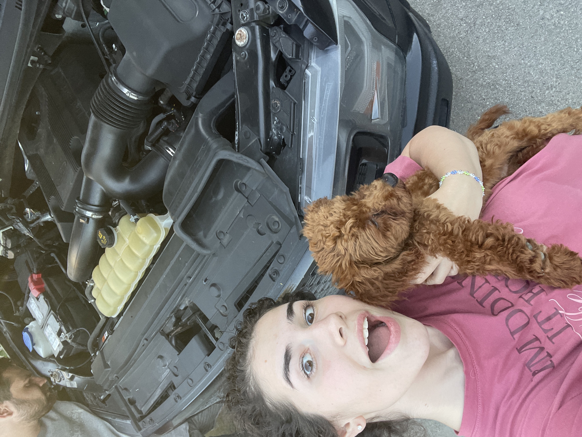 The image shows a woman lying on her back, holding a brown poodle puppy. She has dark, curly hair, fair skin, and is wearing a pink t-shirt. Her mouth is open in an expression of surprise or excitement. The puppy is nestled against her chest. The woman is positioned next to the open hood of a black vehicle. The engine compartment of the vehicle is visible, showing various mechanical components. The background appears to be a concrete or asphalt surface. Location information is not present in the picture.