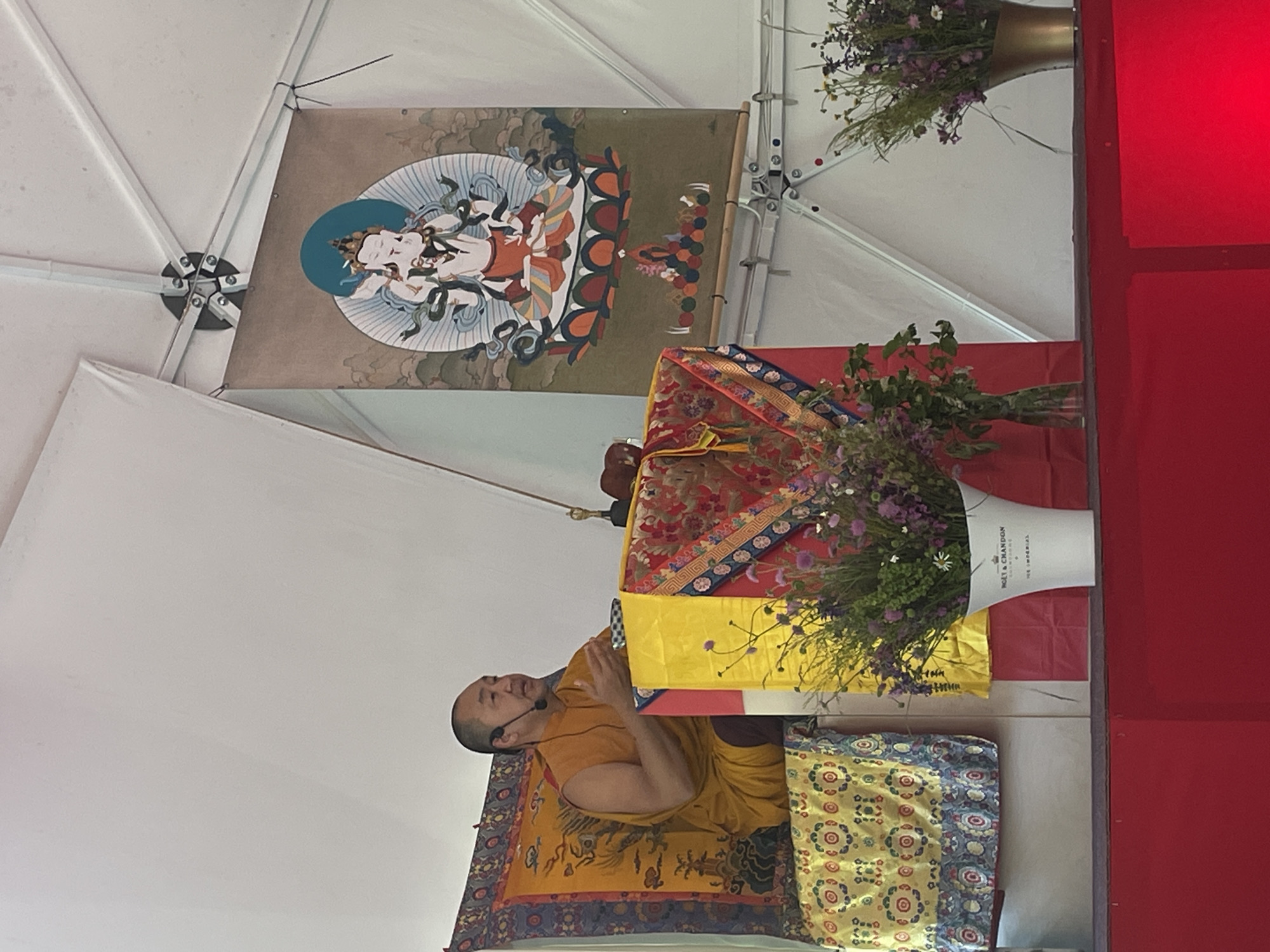 The image shows a monk in a saffron robe, seated on a decorated yellow and blue patterned cushion, wearing a microphone, and speaking. He is positioned in front of a table covered with a red, yellow, and floral-patterned cloth and two floral arrangements. Above him, a banner depicting a deity hangs from the ceiling, and on the right, there's a red wall or partition. The scene appears to be set up under a white tent-like structure. While the specific location within Polja, Montenegro is unknown, the scene suggests a religious or spiritual event.