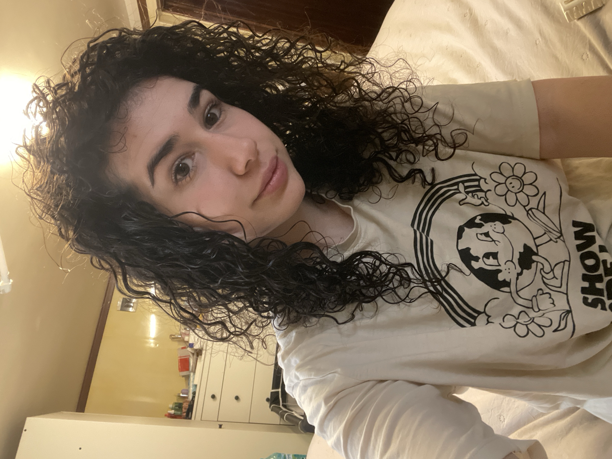 A person with dark, curly hair is lying on a bed with light-colored sheets. They are wearing a light-colored t-shirt with a black and white graphic design that includes a rainbow, a flower, and an animated globe. The person is facing the camera. In the background, there is a dresser with drawers and various items on top. The lighting appears to be indoor, possibly in a bedroom. The image provides no evidence to support a location in Sofia, Bulgaria.