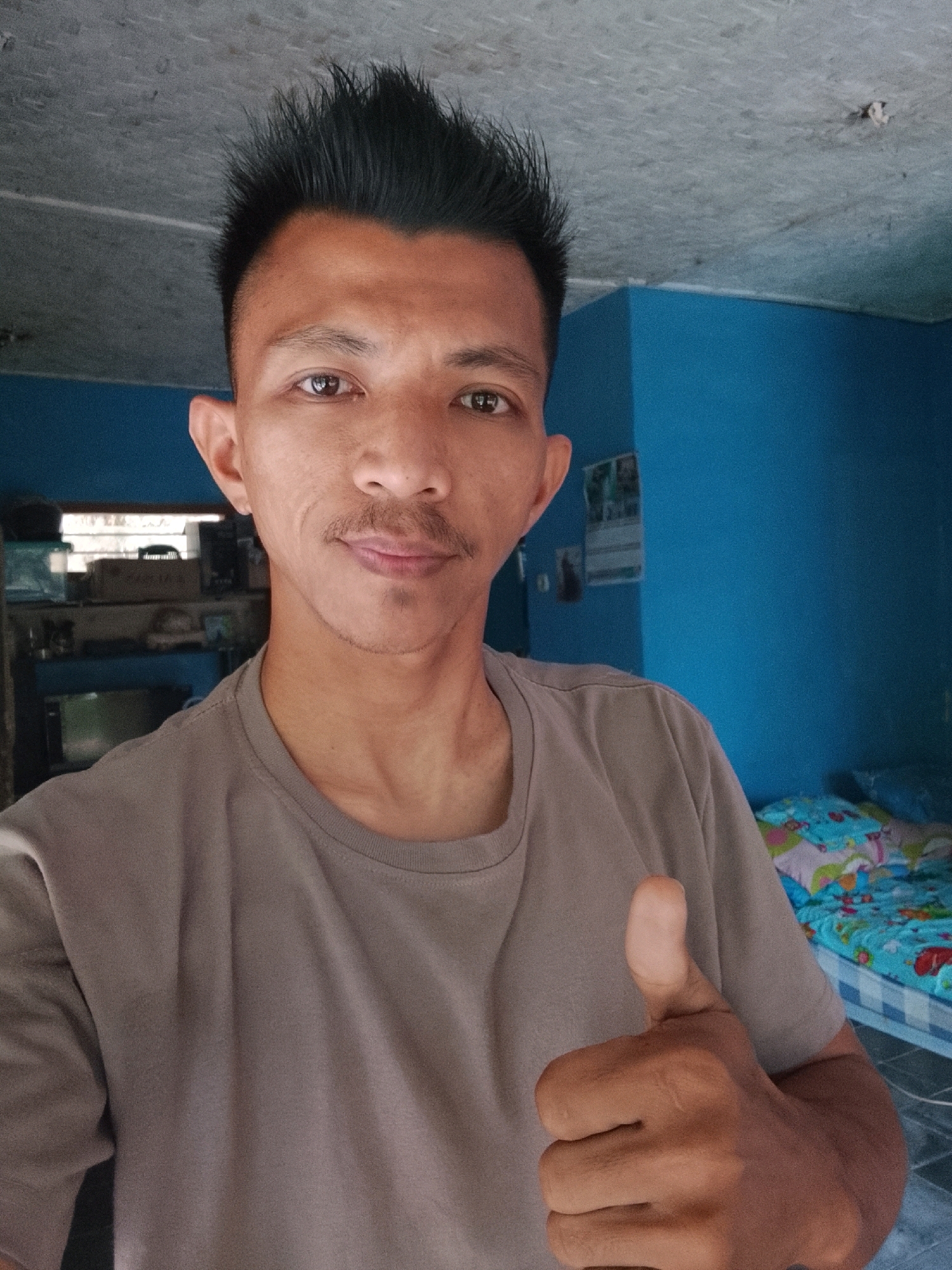 A man with short, spiky black hair and a small mustache is taking a selfie in an indoor setting. He is wearing a taupe t-shirt and is giving a thumbs-up gesture with his right hand. The background shows a blue wall, a ceiling with some marks, and a bed with a colorful patterned comforter. There are also some shelves with items on them visible behind him. The lighting suggests it is daytime, likely in a residential area.