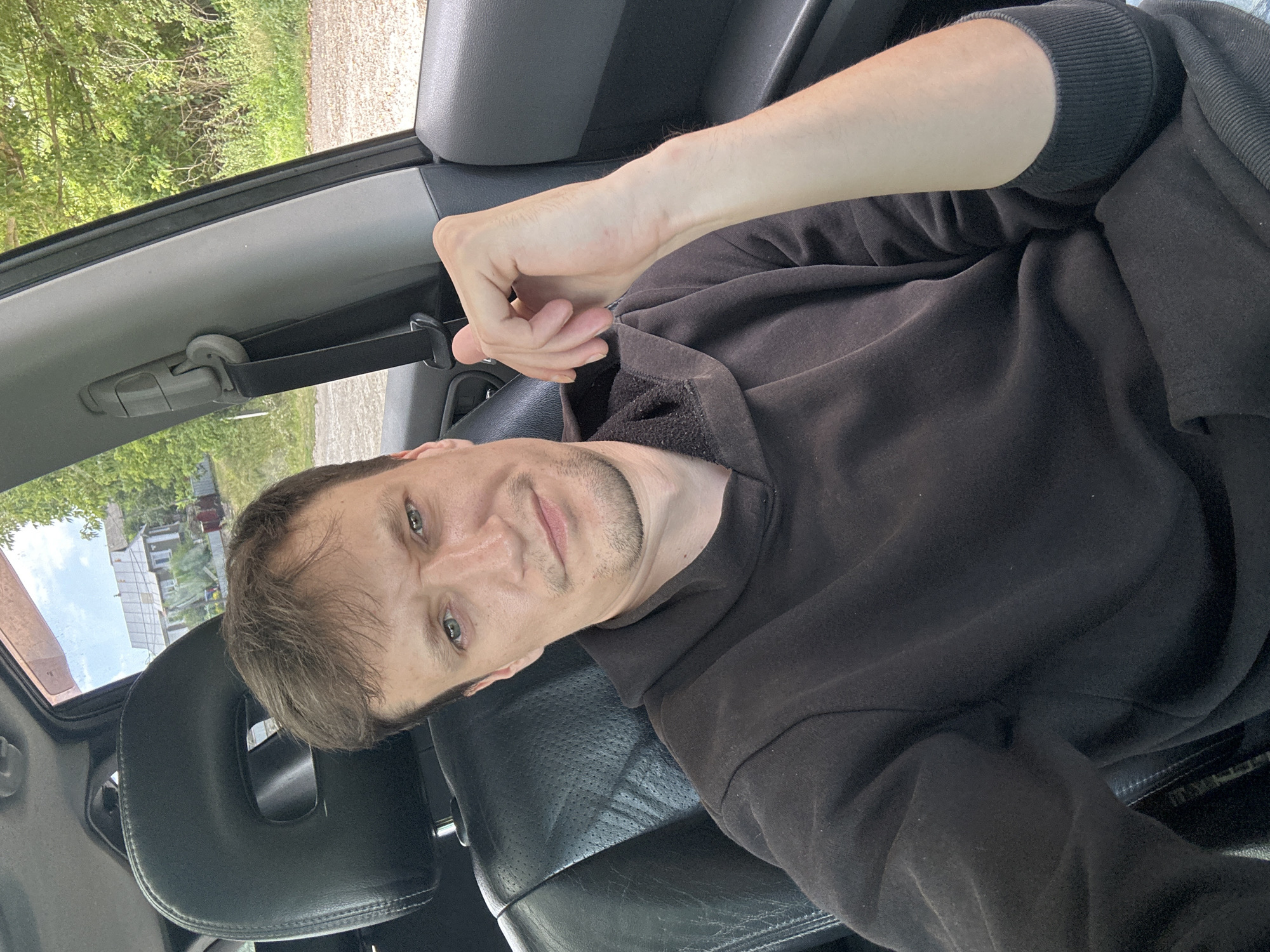 A medium shot shows a man sitting in the driver's seat of a car, looking towards the camera. He has short, brown hair, light skin, and light green eyes. He is wearing a dark brown hooded sweatshirt. His left arm is resting on his lap, and his right hand is on the back of the driver's seat. The interior of the car is visible, including the black leather seats and the headrest. Outside the car window, a rural landscape with trees and houses can be seen. The image was taken in Makiv, Ukraine.