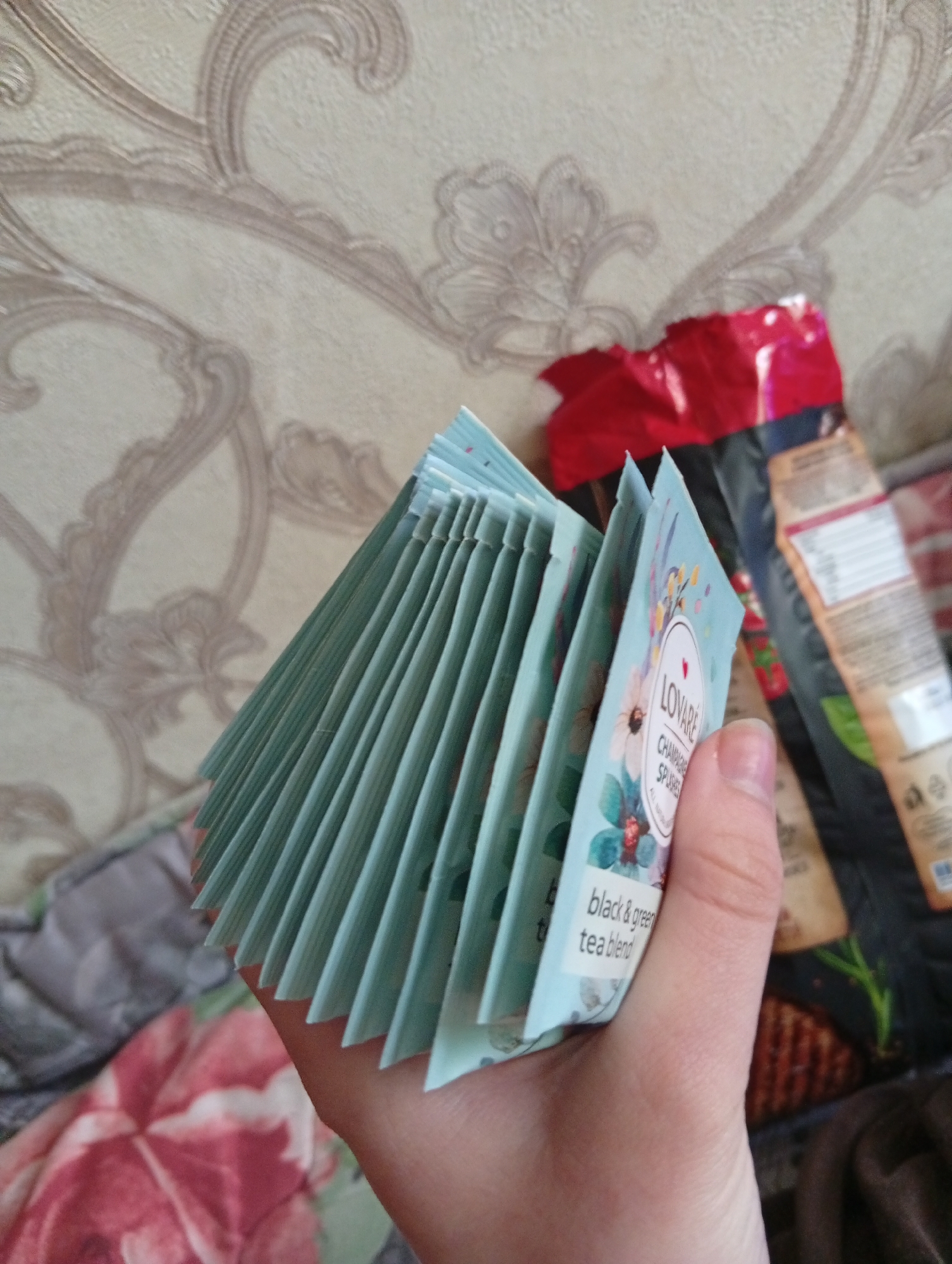 A close-up shot shows a person holding a stack of Lovare "Champagne Splashes" black & green tea blend packets in their hand. In the background, there is a ripped, opened package of a food item. The backdrop includes a patterned wall covering and floral bedding. There is no indication of the location in Poltava, Ukraine.