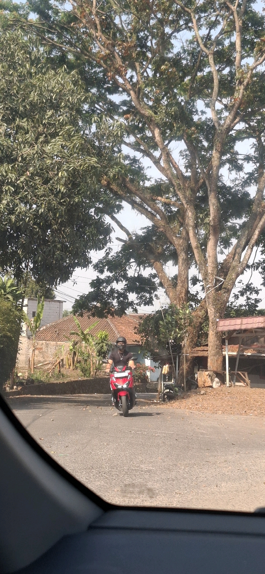 A person wearing a black shirt, black helmet, and a dark mask is riding a red scooter on a paved road. The scooter has a silver emblem on the front and is facing towards the camera. The person is holding the handlebars. The road is a two-lane street with a slight curve. On the right side of the road, there is a wooden structure with a red roof, possibly a stall or shelter, and dry leaves on the ground. Behind the stall, there are trees with thick branches and sparse foliage. On the left side of the road, there are houses with red tile roofs and some banana trees. The background is a clear sky, suggesting daytime. The scene appears to be in a semi-rural or suburban area of Cibinong Hilir, Indonesia. The image is captured from inside a vehicle, indicated by the blurred dark foreground which resembles a car's interior.