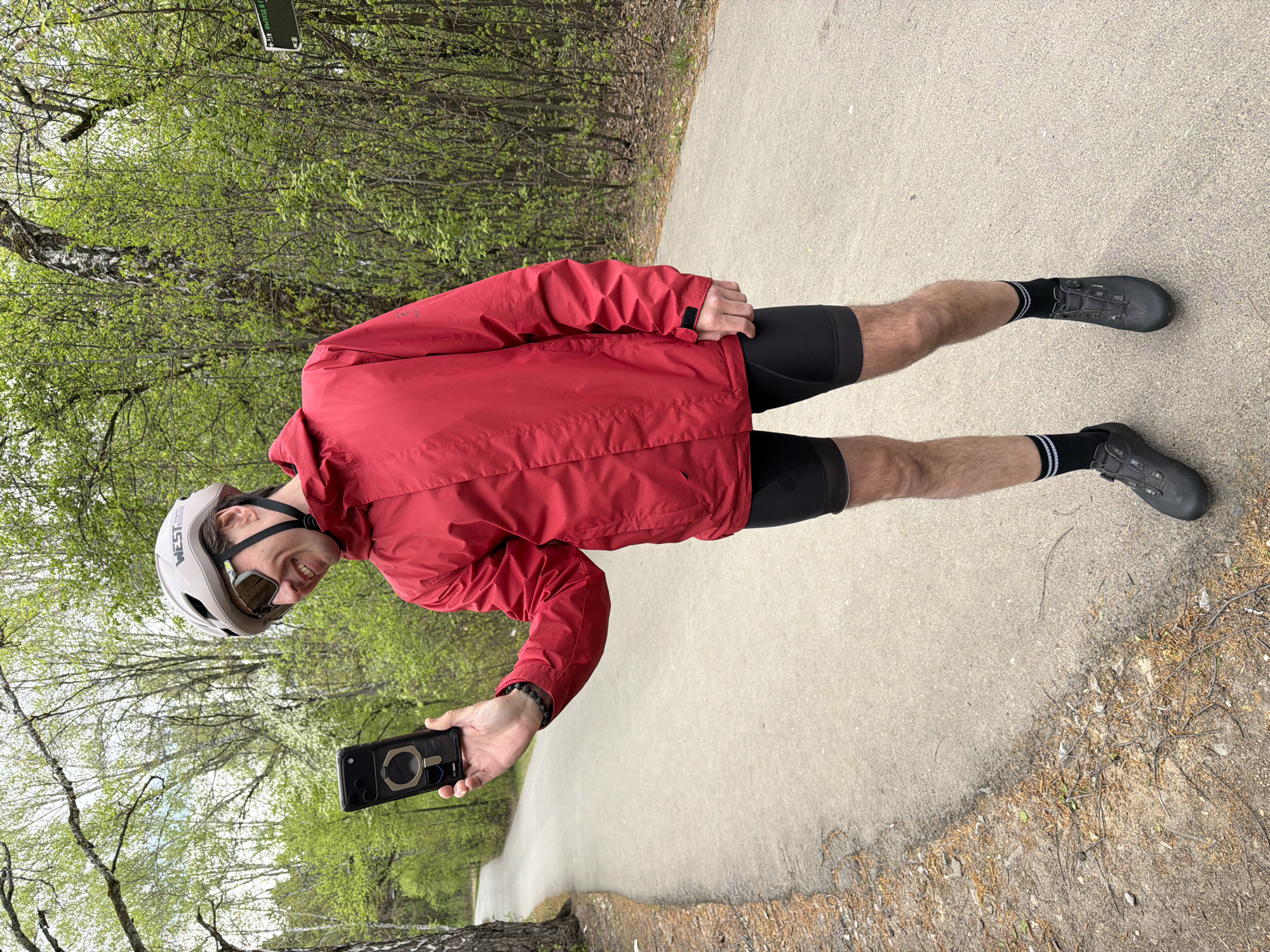 A cyclist stands on a paved path holding a smartphone. The cyclist wears a red jacket, black cycling shorts, black socks with white stripes, and grey cycling shoes. The cyclist also wears a grey helmet and sunglasses. The background includes trees with green leaves on either side of the path. The path appears to be a pedestrian or cycling path within a park-like setting. Based on the foliage and the cyclist’s clothing, the season appears to be spring or early summer. The location is likely in or near Tyumen, Russia.