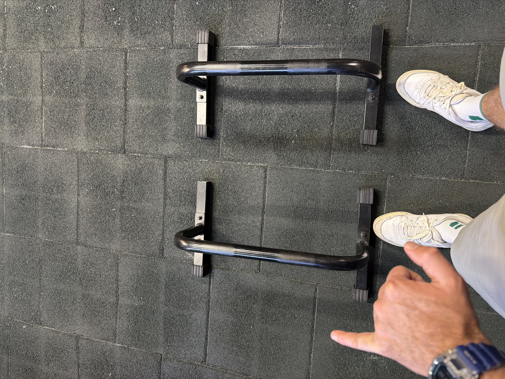 The image depicts an indoor scene on a grey rubber-tiled floor. Two sets of black, parallel dip bars are present. A person is visible with white sneakers, grey pants, and a watch. The person's hand is making a "hang loose" gesture. The location is not identifiable as being in Tbilisi, Georgia from the visual data.
