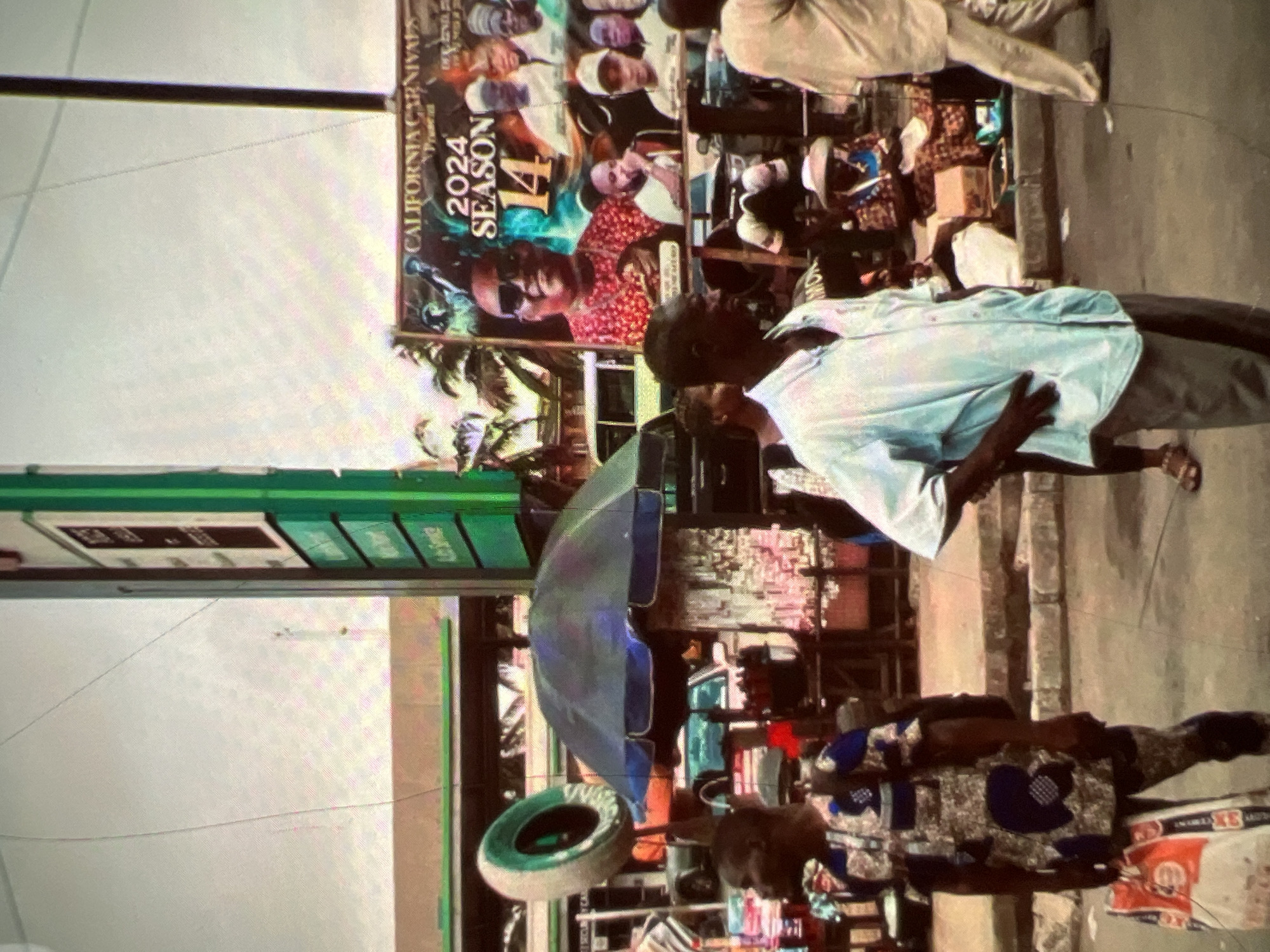 The image depicts a street scene in Lagos, Nigeria. In the foreground, a man wearing a patterned traditional Nigerian outfit stands with his left hand on his hip, carrying a white and orange shopping bag. To his right, another man in a light blue, short-sleeved shirt and grey trousers is also standing with his hands on his hips. Behind them, a large billboard advertises "CALIFORNIA CARNIVALS Present 2024 SEASON 14," featuring portraits of several people. To the left of the billboard and behind the first man, an umbrella shades a stall where goods are displayed. A large tire is mounted on a pole in the midground. A gas station structure with green signage is visible in the background. Several other individuals and vehicles are present in the scene, suggesting a busy urban environment.