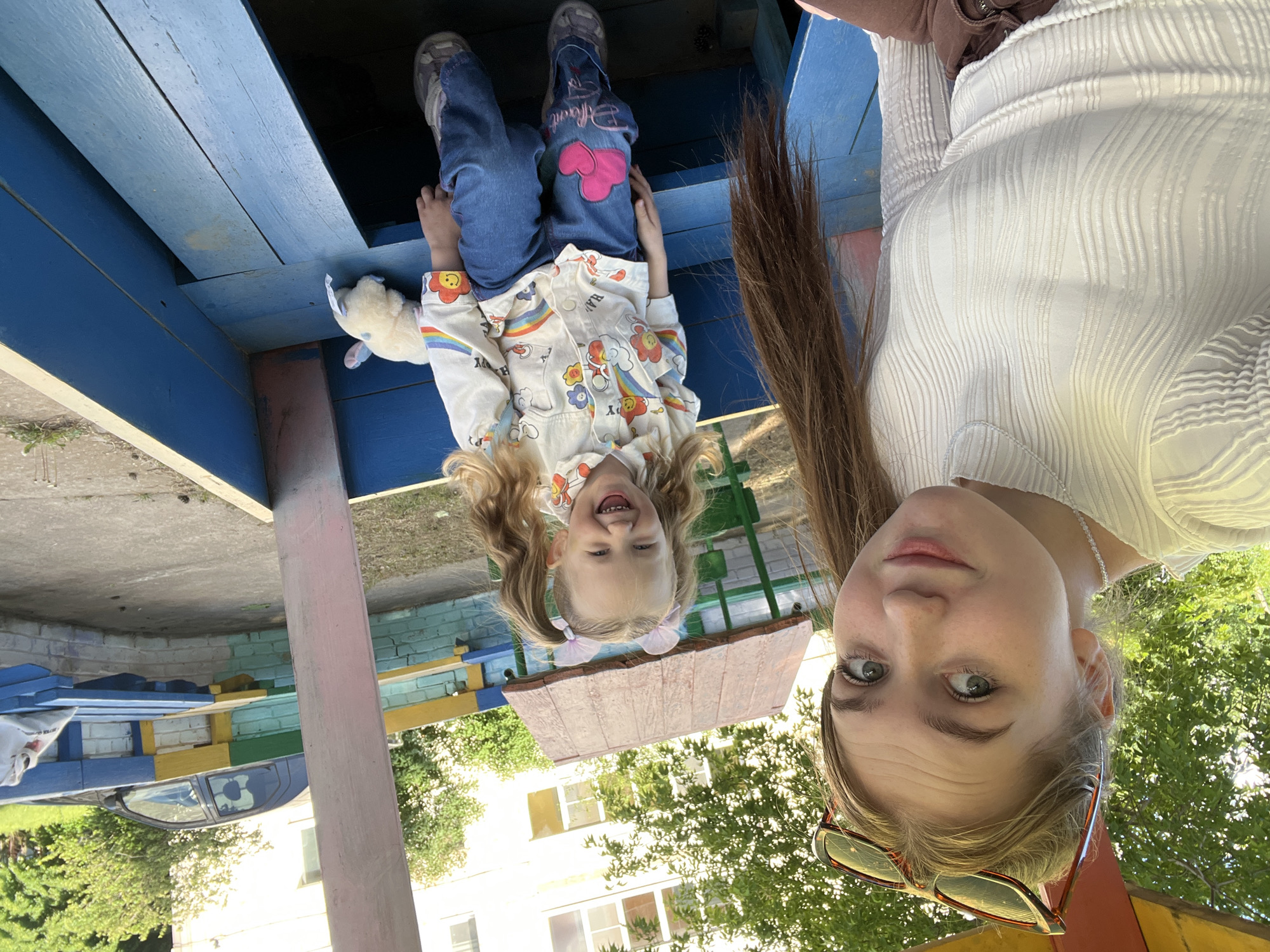 The image shows a selfie taken by a fair-skinned woman with shoulder-length brown hair, green eyes, and sunglasses resting near her forehead. She is wearing a white textured blouse and a pearl necklace. Beside her, a young girl with blonde hair in pigtails, adorned with a pink hair accessory, is hanging upside down. The girl is wearing a rainbow-themed shirt and jeans with a pink heart patch. They appear to be under a raised wooden structure, part of a children’s playground. A plush toy is also visible within the structure.
The scene appears to be outdoors, with a building and greenery visible in the background, and a car can be seen reflected in a metallic surface. The image was taken in the city of Chaikovsky, Russia.