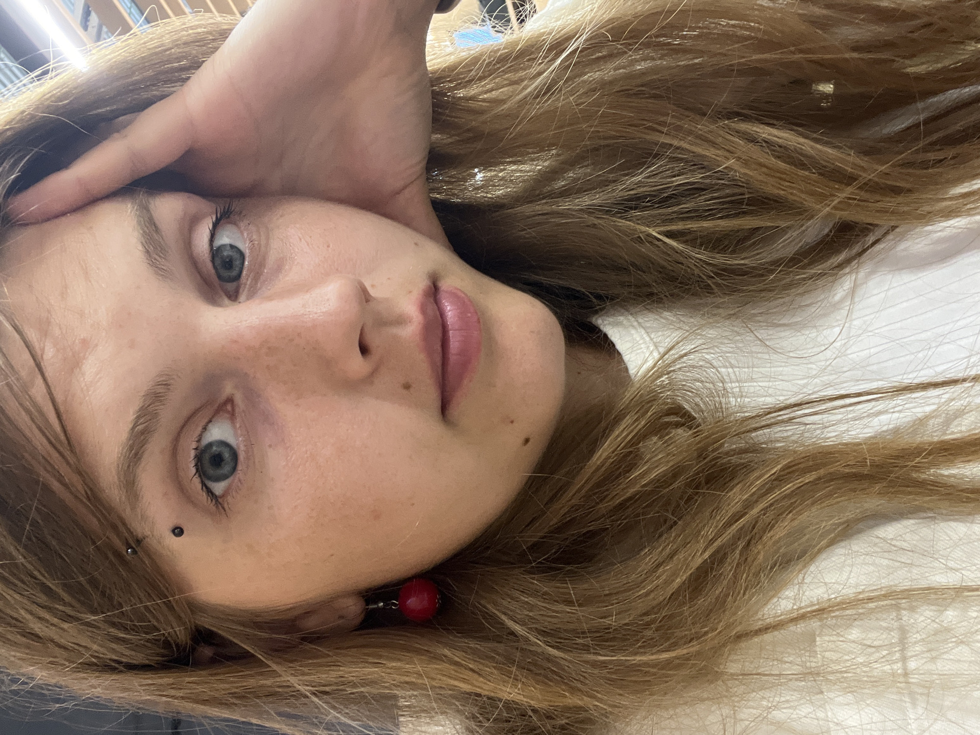 A close-up, head-and-shoulders portrait shows a young woman with light brown, shoulder-length hair, a light complexion with freckles, and blue eyes. She has a vertical eyebrow piercing with two black balls. A red, spherical earring hangs from her left ear. Her right hand is propped on her forehead. She is wearing a white, ribbed t-shirt. The background is out of focus and shows a ceiling with strip lighting. There is no information available to determine the location in the city of Minsk, Belarus.