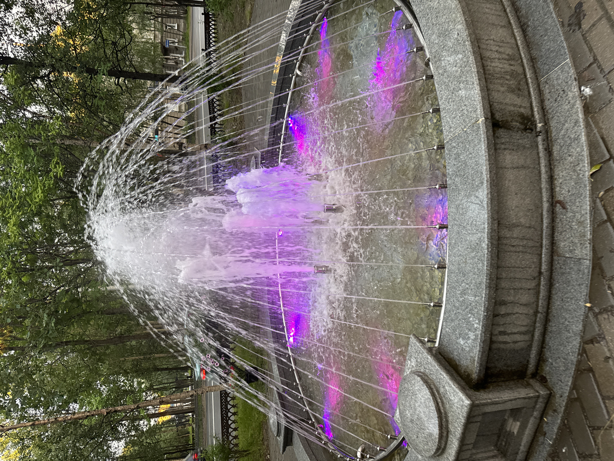 A fountain in a park is illuminated with purple lights, creating a vibrant display against the cascading water. The fountain is made of granite and has a metal rim around the basin. The water shoots upwards in a central spray and also emerges from smaller nozzles around the perimeter, creating a delicate, curtain-like effect. In the background, trees line a street where a few cars are visible. The ground around the fountain is paved with brick. The sky is overcast and the lighting suggests it is either early morning or late evening.