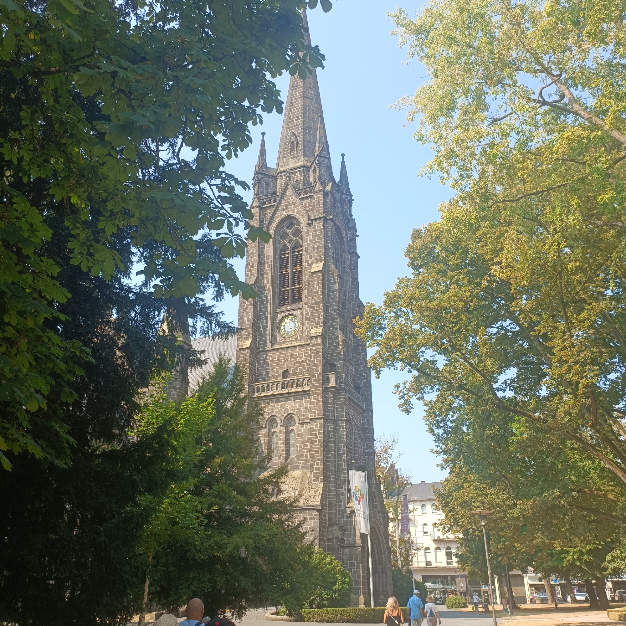 The image shows a view of the Johanneskirche in Bad Nauheim, Germany, from the northeast. The church's stone tower, topped with a spire, is visible behind lush green trees. The clock on the tower reads approximately 2:45. In the foreground, several people are walking on a paved path. A white banner with a colorful logo hangs from the church. The sky is clear and blue, indicating a sunny day. Buildings are visible in the background, suggesting an urban setting.