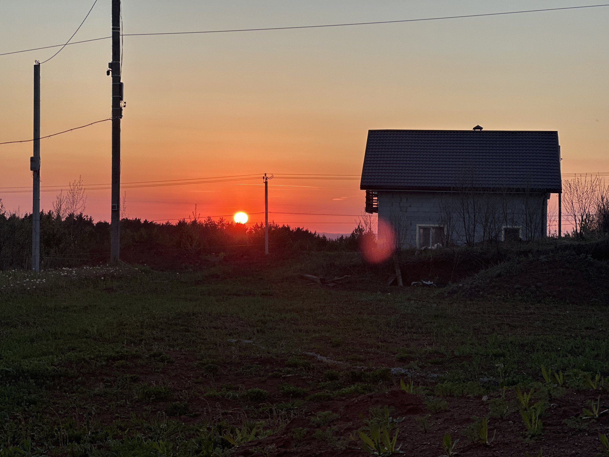 The image depicts a rural landscape at sunset in or near Прикамский, Russia. In the foreground, a grassy field transitions to a dirt area with sparse vegetation. Two weathered utility poles stand on the left, with a third, equipped with electrical boxes, closer to the center. A small, unadorned house with a dark gray roof is located to the right. The setting sun, a bright orange disk, is visible on the horizon through a line of trees. The sky fades from orange near the horizon to a pale gray-blue overhead. There are no visible people in the image.