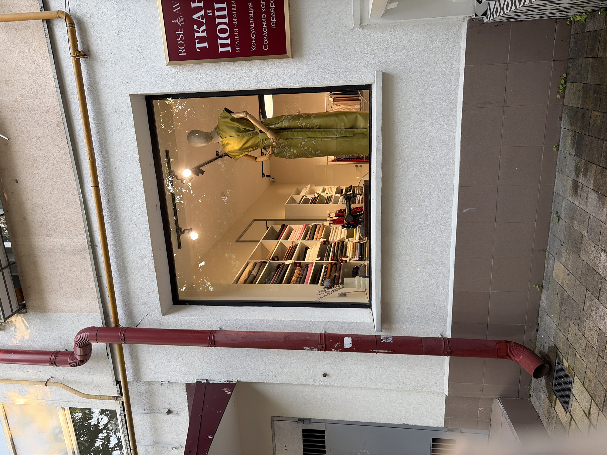 The image shows the exterior of a shop with a large display window. The shop appears to be a fabric store, as rolls of fabric are neatly displayed on shelves visible through the window. A mannequin is positioned in the window, wearing a long, greenish-yellow dress. To the right of the display window, a red sign with white text is mounted on the wall. The building has a textured white facade, with a dark brown tiled base. A red drainpipe runs vertically along the left side of the building, with several bends. Yellow pipes are visible running horizontally along the upper part of the building. The ground in front of the shop is paved with square grey tiles, and a drain grate is visible. Small green plants are growing in cracks in the paving near the base of the building. The scene is outdoors, with hints of greenery reflected in the display window, suggesting trees or foliage nearby. The overall impression is of a retail establishment in an urban environment.