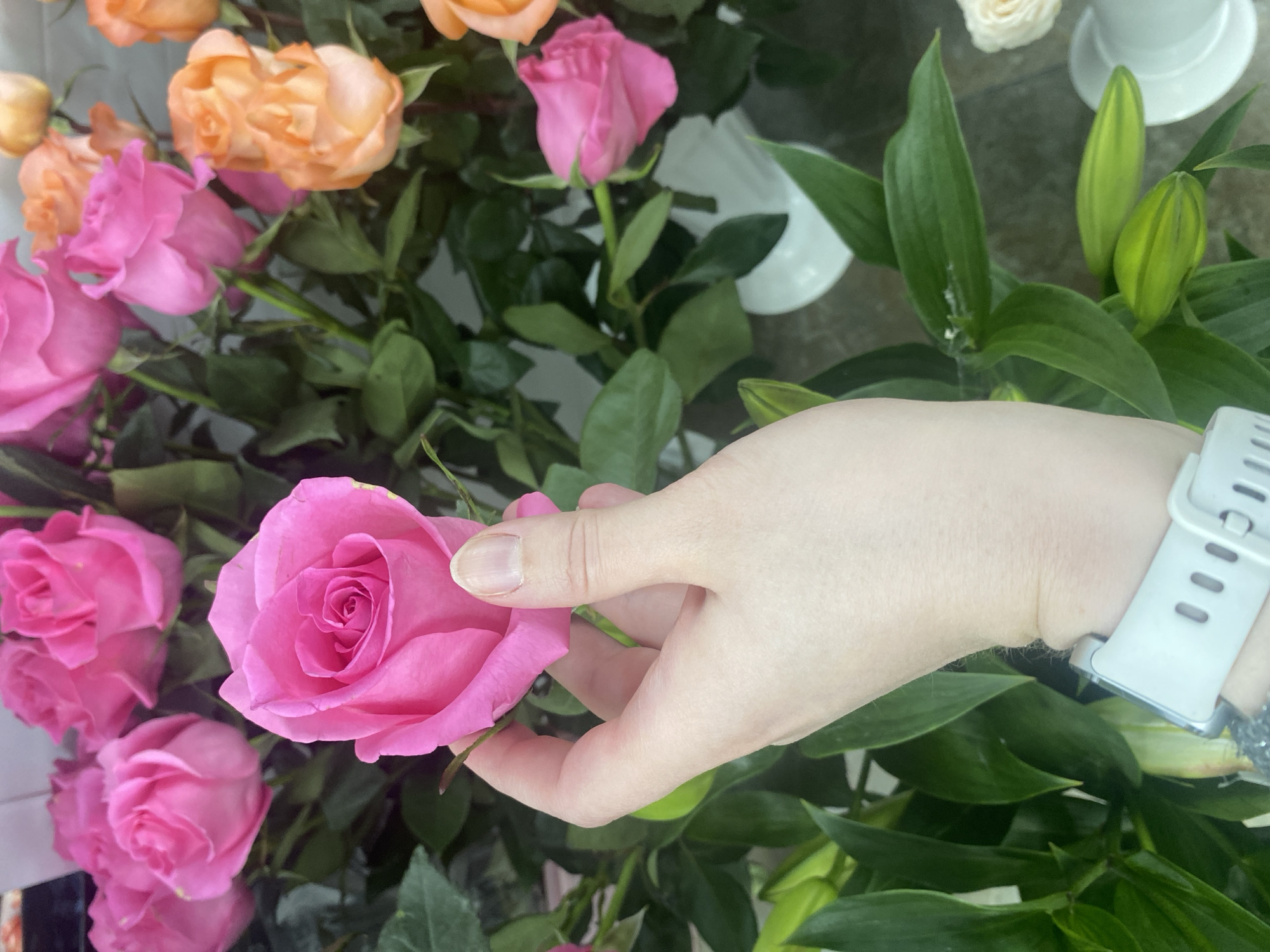 A hand is holding a bright pink rose among a bouquet of pink and orange roses, as well as green lily leaves with unopened buds. The background appears to be an indoor setting, potentially a flower shop or a home. There is no information available to place the location in Yekaterinburg, Russia.