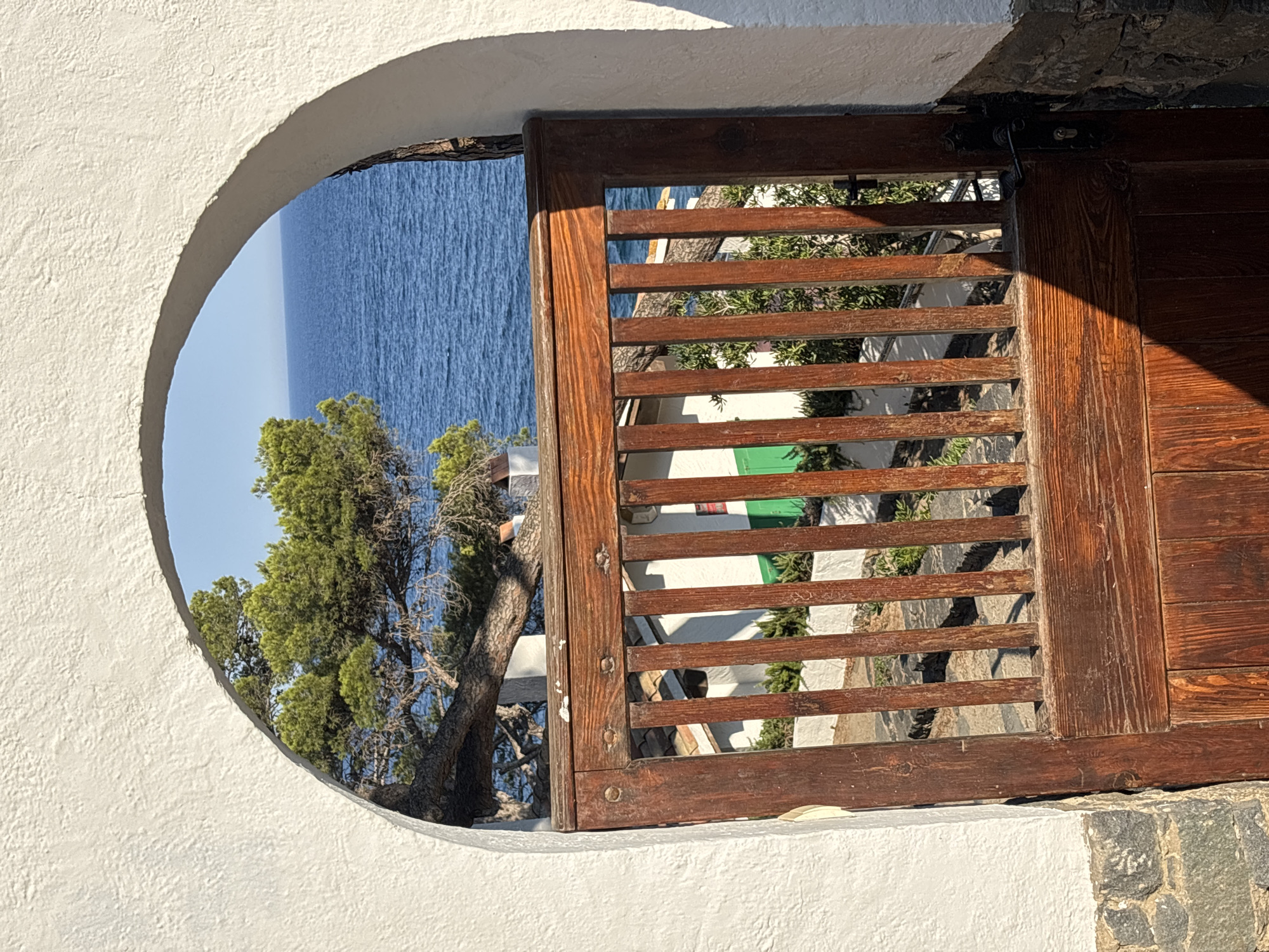 An arched white wall frames a view of the Mediterranean Sea and a pine tree through a wooden gate. The gate is constructed of vertical wooden slats and has a dark metal latch. In the background, a white building with a green shutter is partially visible. The scene is set in Calella De Palafrugell, Spain, a coastal town on the Costa Brava. The sunlight casts a strong shadow across the gate.
